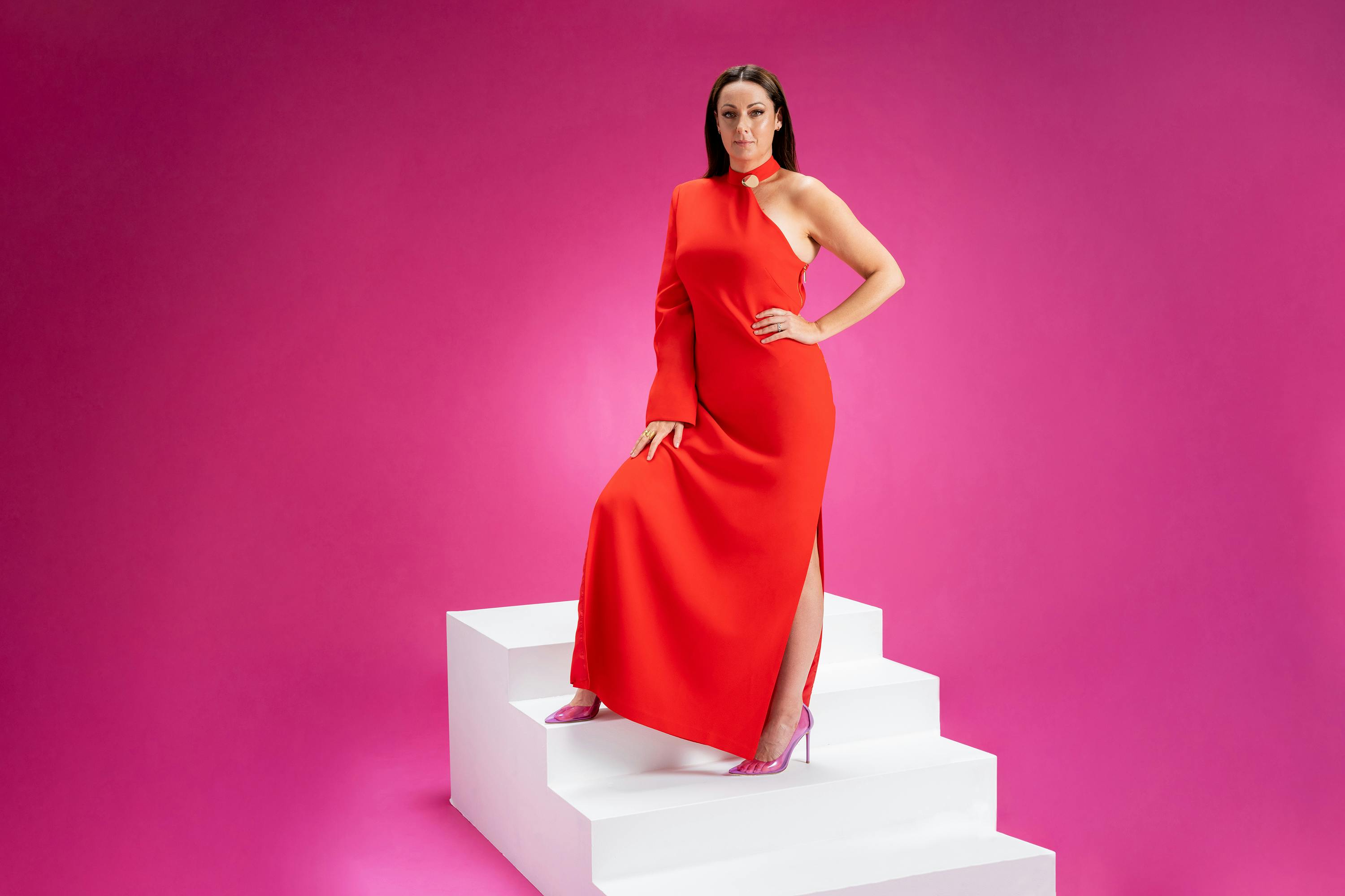 Celeste Barber wears a long red dress and poses on a staircase against a pink background.