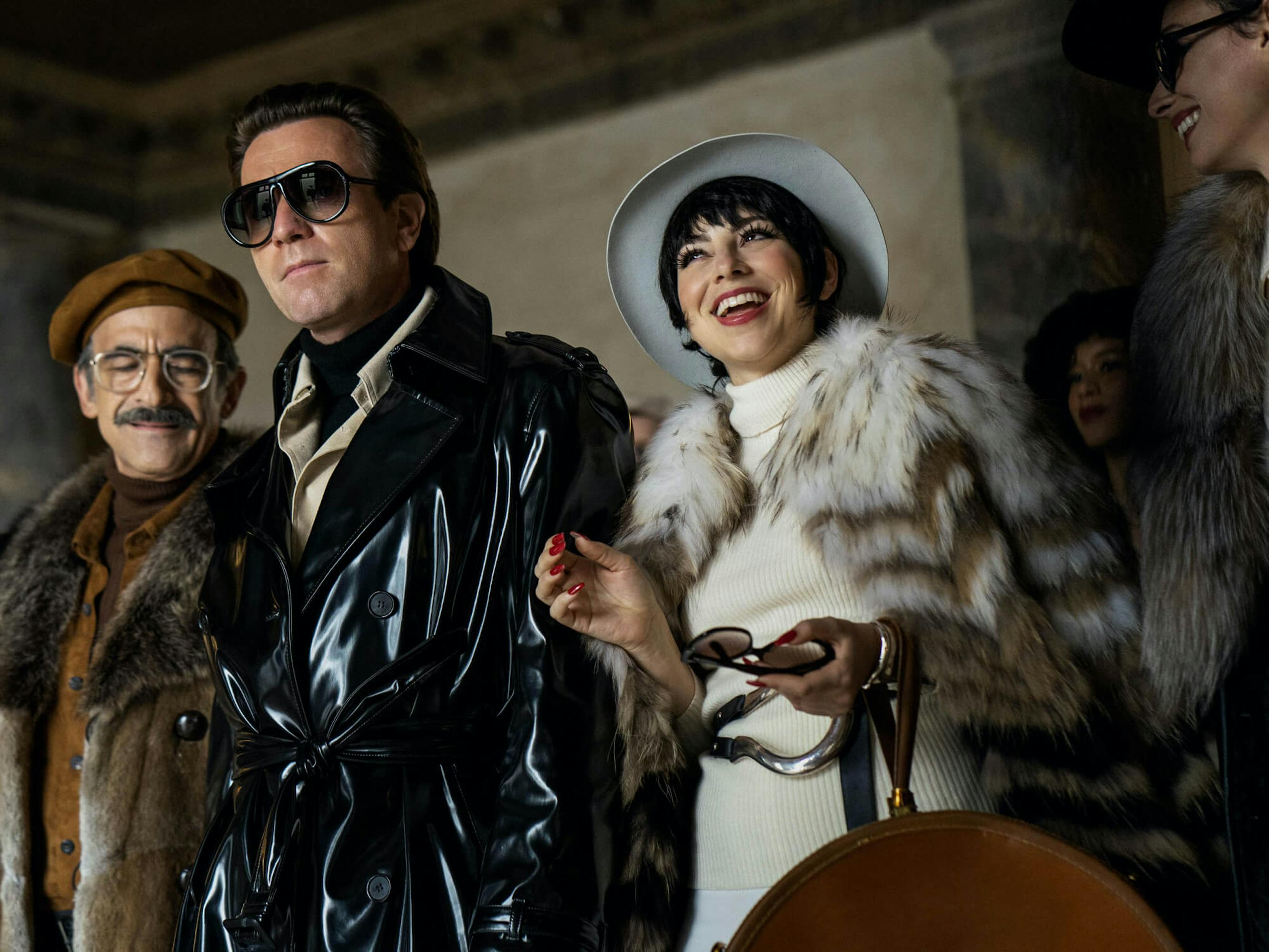Joe Eula (David Pittu), Halston (Ewan McGregor), and Liza Minnelli (Krysta Rodriguez) stand in a line wearing brown, black, and white fur coats respectively.