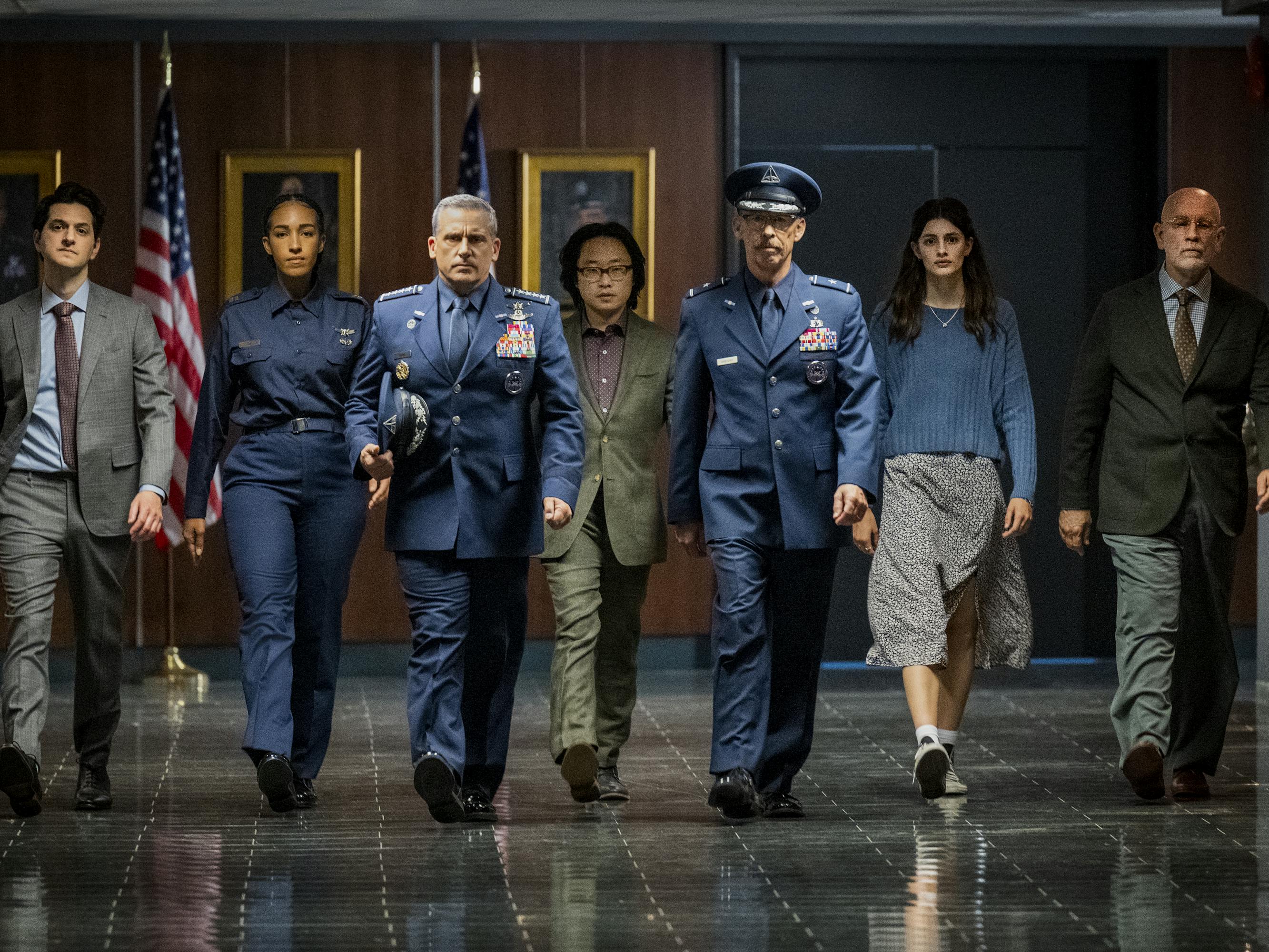 Ben Schwartz, Tawny Newsome, Steve Carell, Jimmy O. Yang, Don Lake, Diana Silvers, and John Malkovich walk down an empty hallway. Some of the men are wearing uniforms, and they all look serious. 