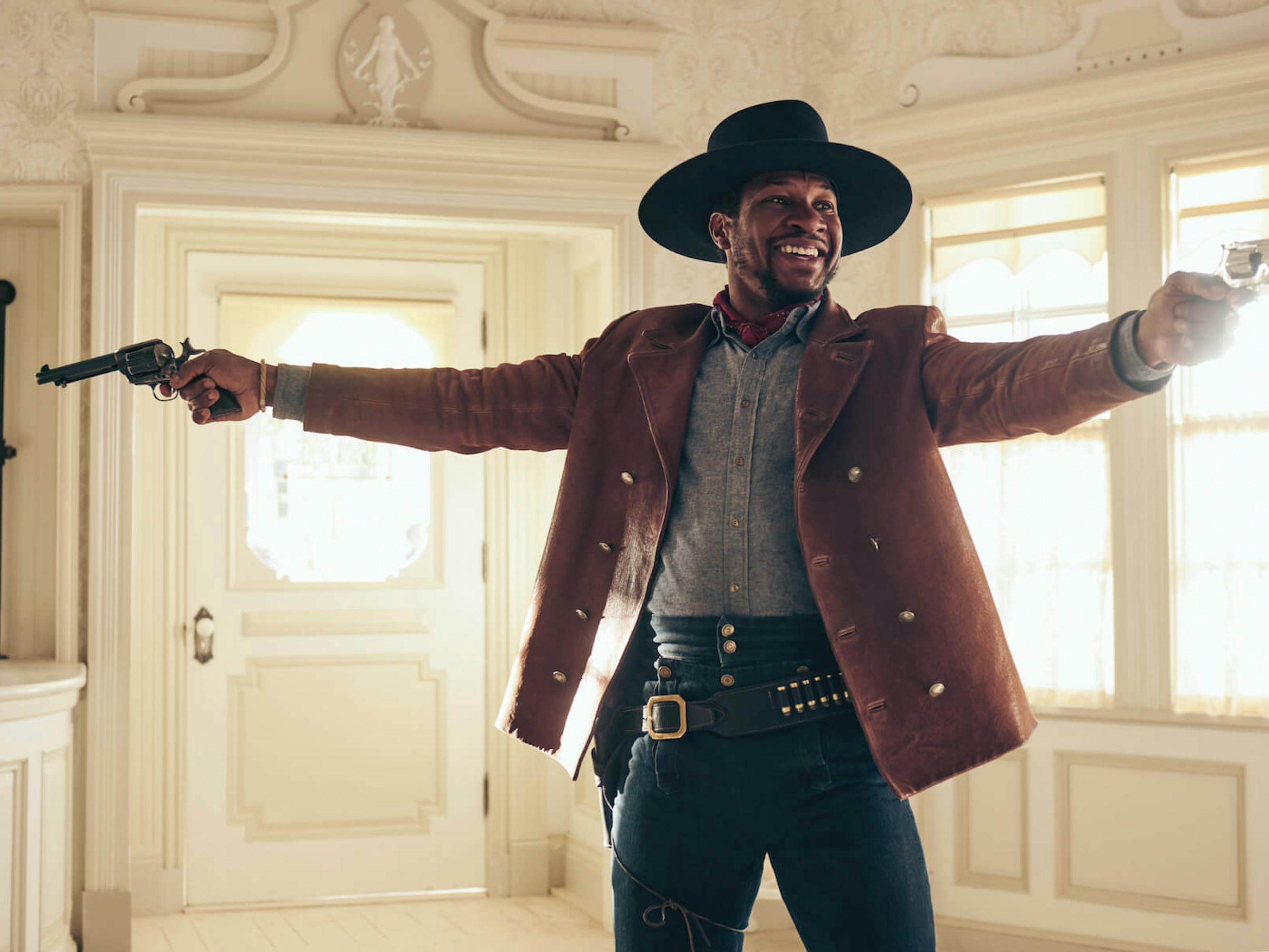 Nat Love (Jonathan Majors) holds two guns in opposing directions. He wears a dark hat, gray shirt, brown jacket with gold peacoat buttons, a red handkerchief, and navy pants. His smile is dissonant with this high intensity picture.