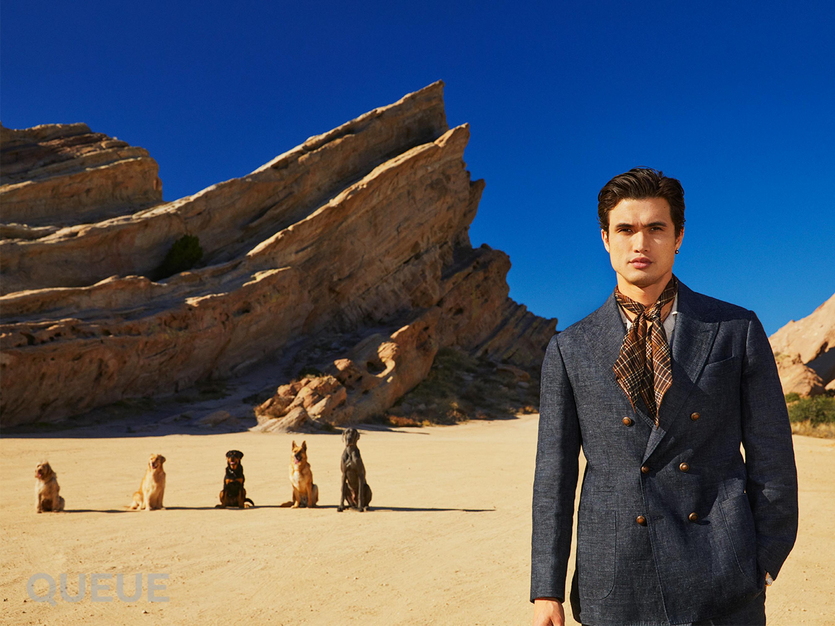 Charles Melton wears a smart suit and neck tie while standing in front of a breathtaking rock formation at the Vasquez Rocks Natural Area and Nature Center in Agua Dulce, California; an obedient group of dogs sit in a single file line behind him from shortest to tallest, mirroring the shape of the rock formation.
