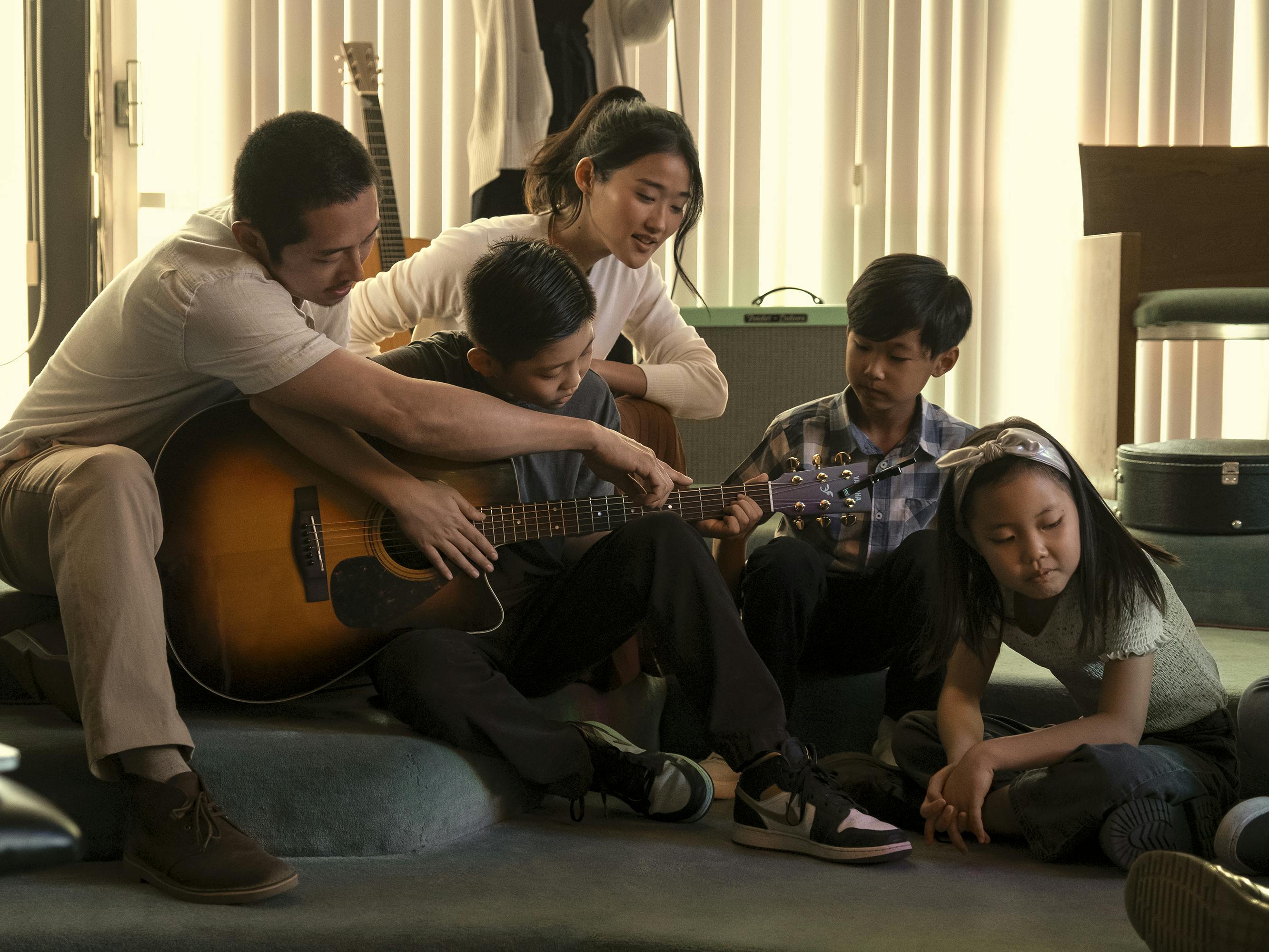 Danny (Steven Yeun) and Esther (Andie Ju) sit with some younger kids at the church around a guitar.