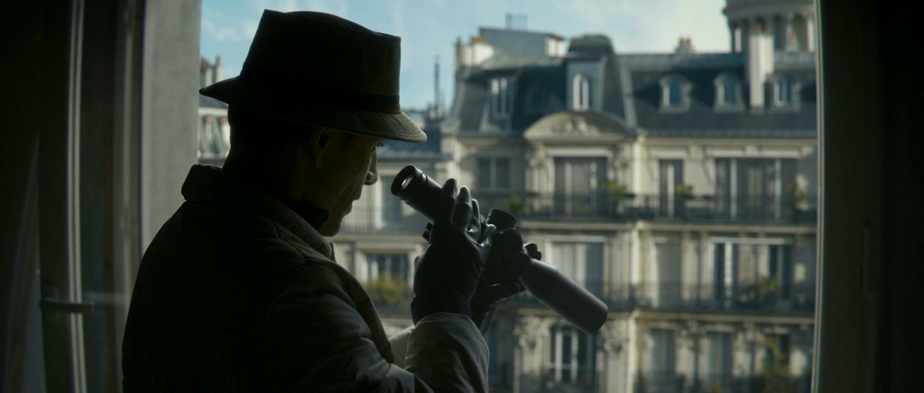 The Killer (Michael Fassbender) looks through a scope and wears a trench coat and a bucket hat.