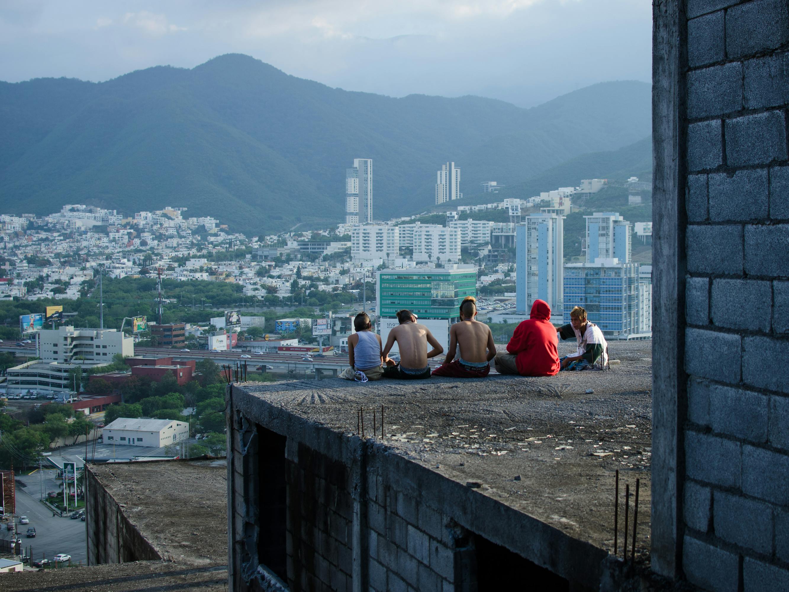 Characters from Ya no estoy aquí sit on the edge of a building overlooking a city.