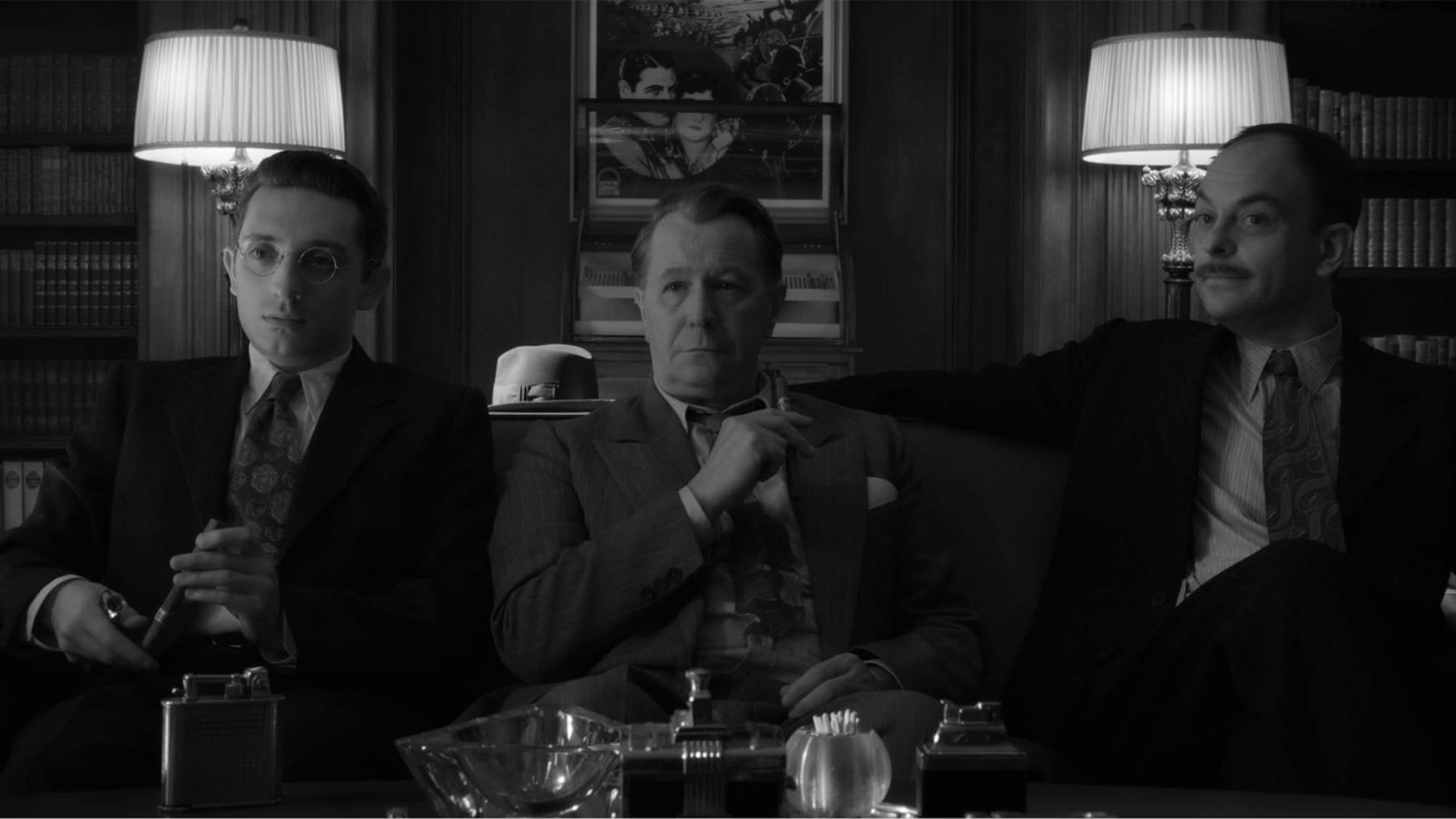In a shot from the film, we see how the scene above translates to a black-and-white experience. Romano, Oldman, and Hecht are pictured on one of the office couches, framed by two of the office’s three lamps.