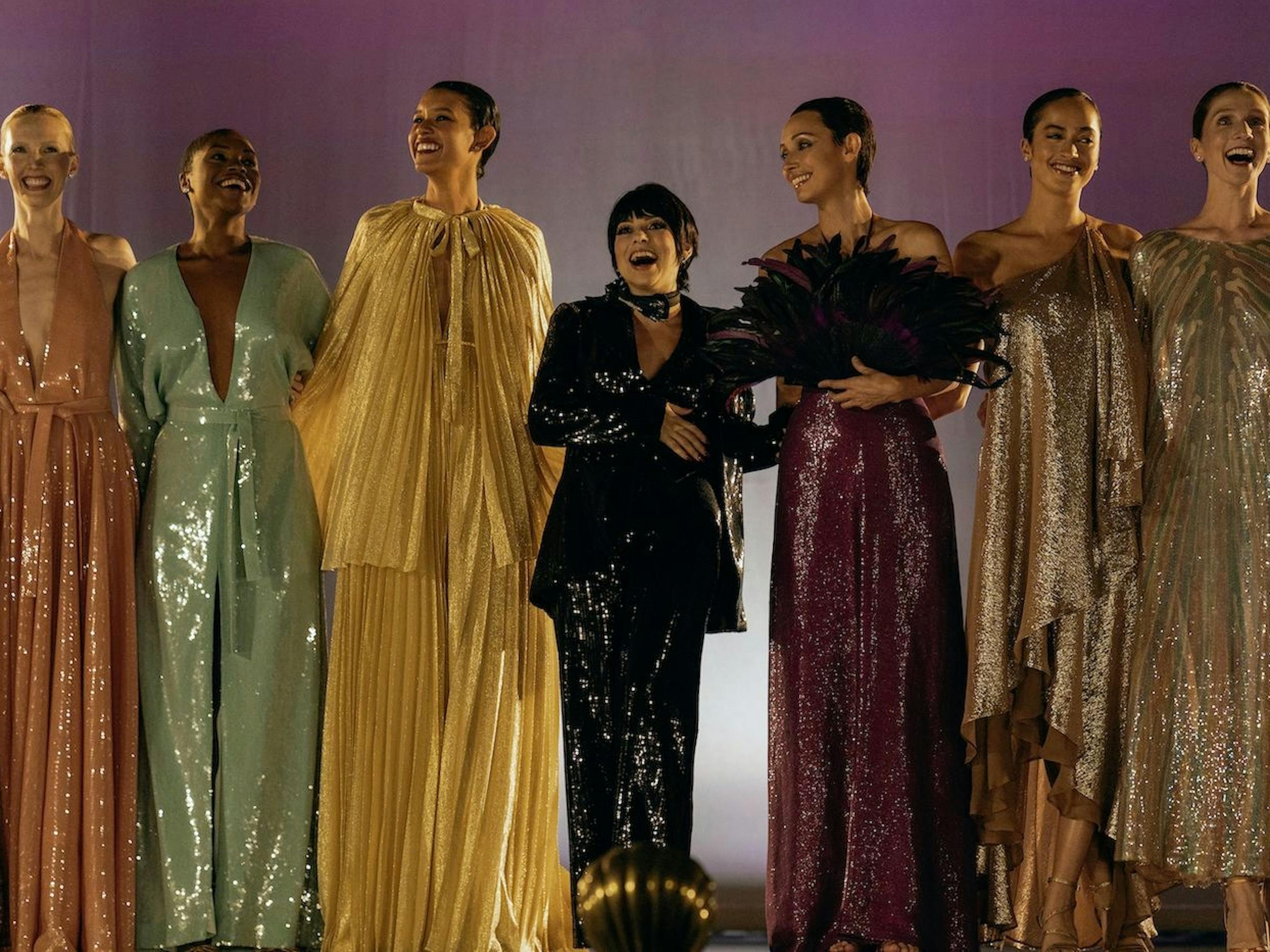 A row of models wear beautiful shimmery gowns. From left to right, there is pink, pistachio green, yellow, black, purple, and metallic gold. Minnelli stands in the middle, in black, next to Peretti in purple. They smile, presumably at the end of the show.