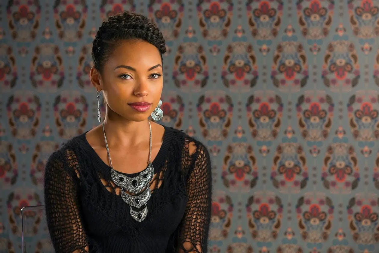 Sam White (Logan Browning) sits in front of an abstract patterned background.  She is wearing a black loose knit top, chunky silver and onyx necklace, and large silver drop earrings. She gazes into the camera with a knowing look. 