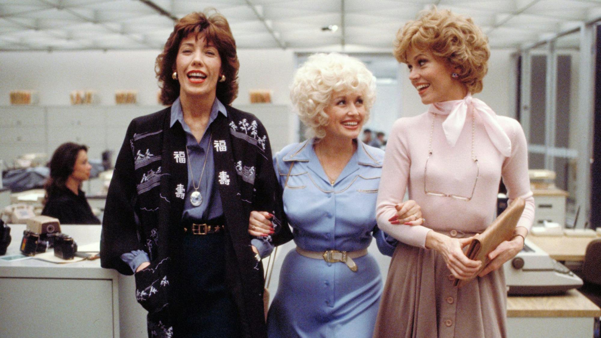 Lily Tomlin, Dolly Parton, and Jane Fonda walk through the office in a still from 9 to 5. Tomlin has on a black jacket over a blue button-up and black trousers. Parton wears a blue shirt dress cinched at the waist with a belt. Fonda wears a pink long sleeved top with a scarf tied around her neck and a high waisted skirt. 