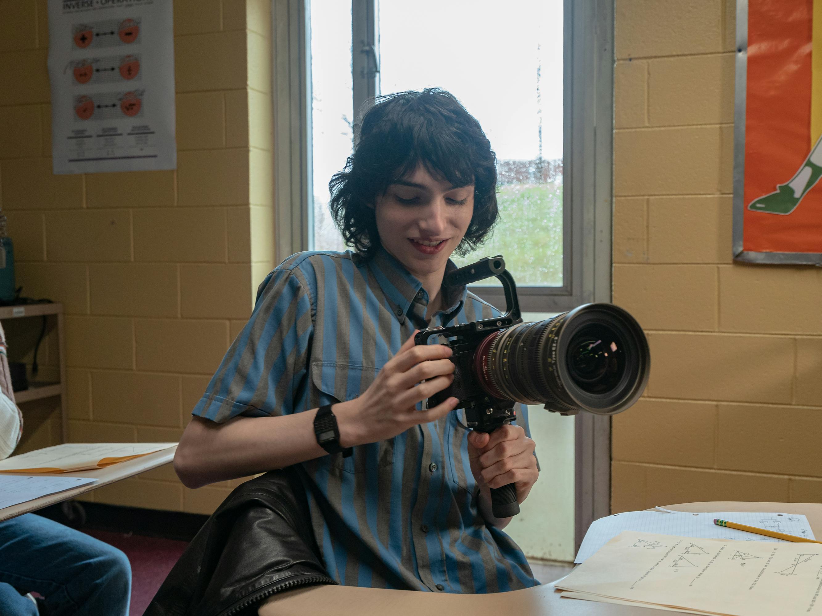 Mike Wheeler (Finn Wolfhard) wears a blue shirt and holds a huge camera on his desk.