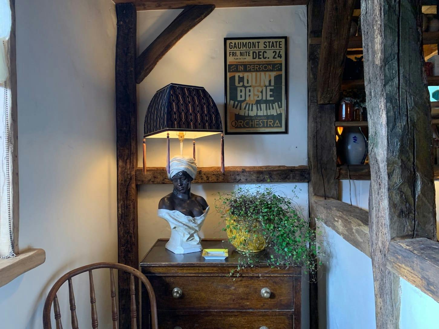 A wooden bureau, with a bust, plant, and lamp atop. There is also a poster on the wall, framed by wood beams throughout this little nook. We can also glimpse the top of a wooden chair.