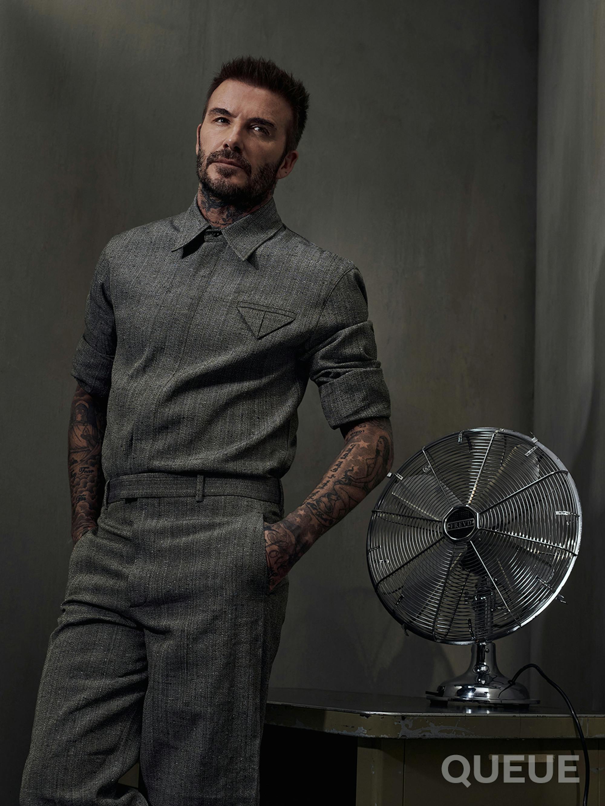 David Beckham wears all gray and stands next to a fan in a gray-walled room. 