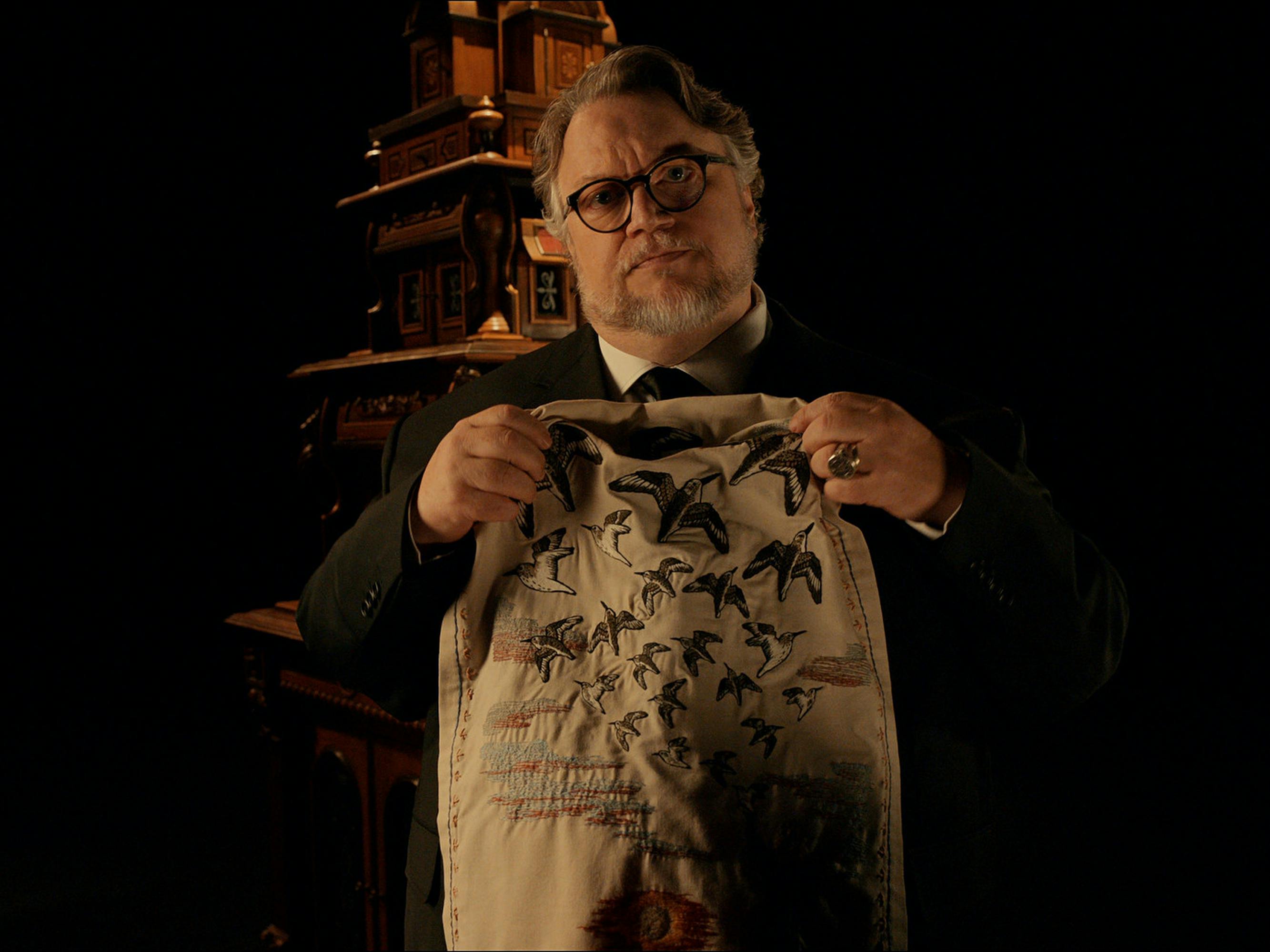 Guillermo del Toro at the start of one of his anthology episodes. He holds a mysterious cloth, decorated with birds, up against his suit. 