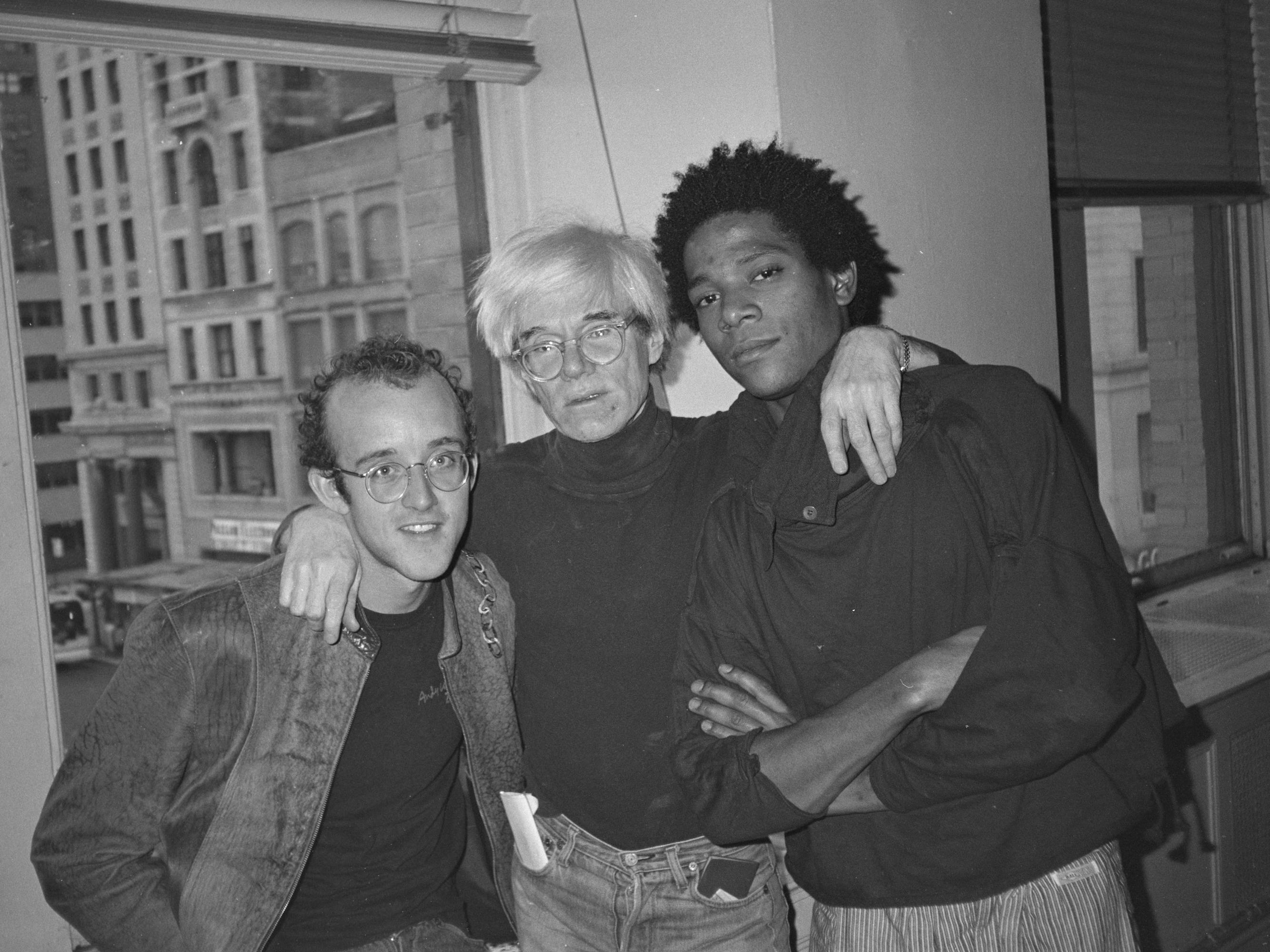 Keith Haring, Andy Warhol, and Jean-Michel Basquiat stand together by a big window. They each wear black tops.