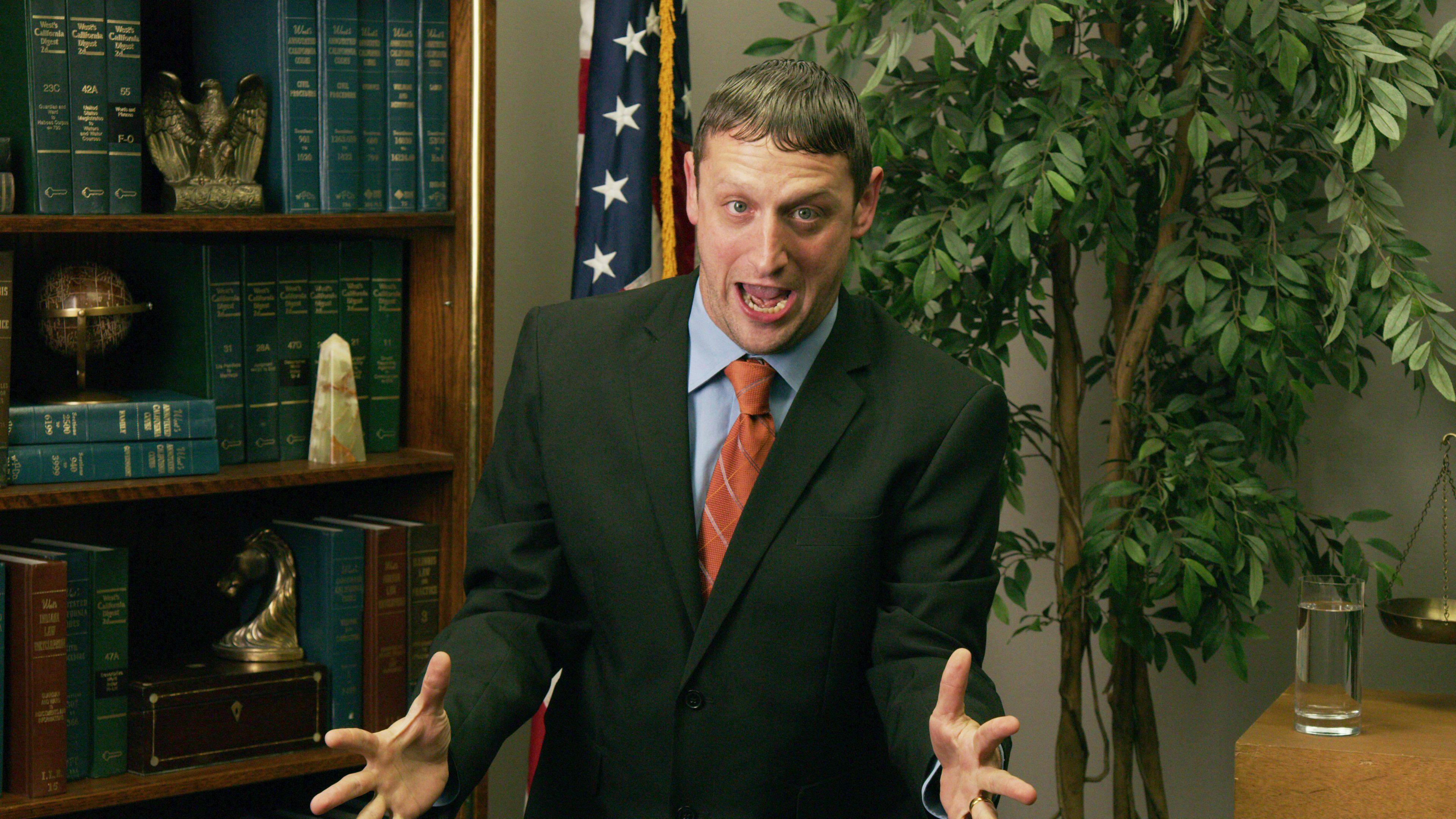 Tim Robinson wears a black suit jacket, blue shirt, and red tie. Behind him is an American flag, plant, and some very legal looking books.