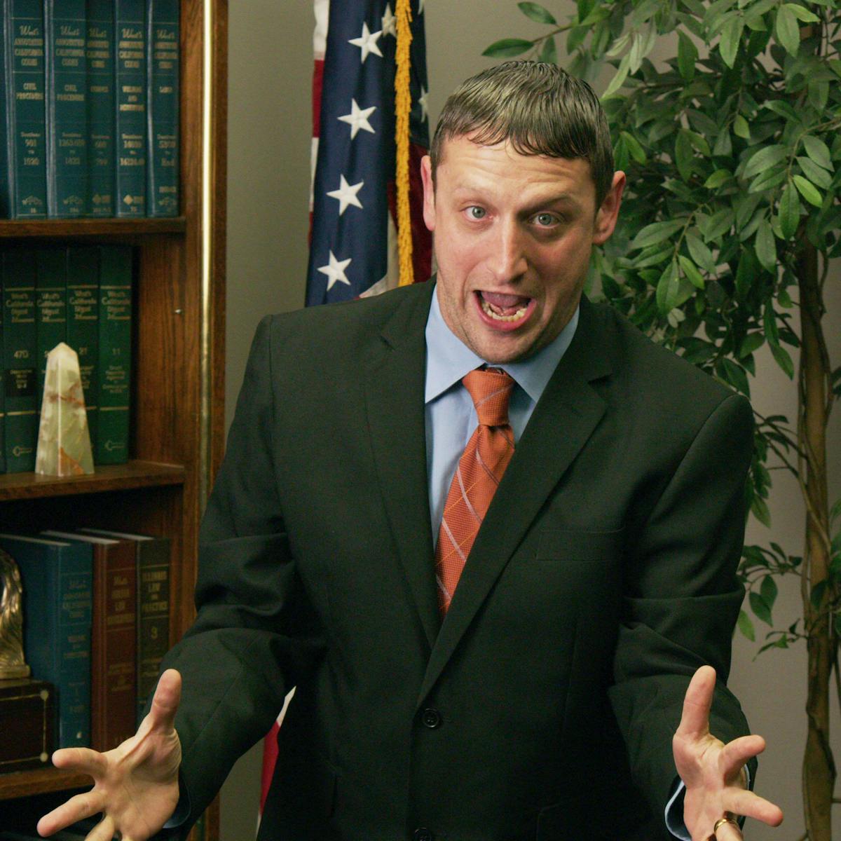 Tim Robinson wears a black suit jacket, blue shirt, and red tie. Behind him is an American flag, plant, and some very legal looking books.