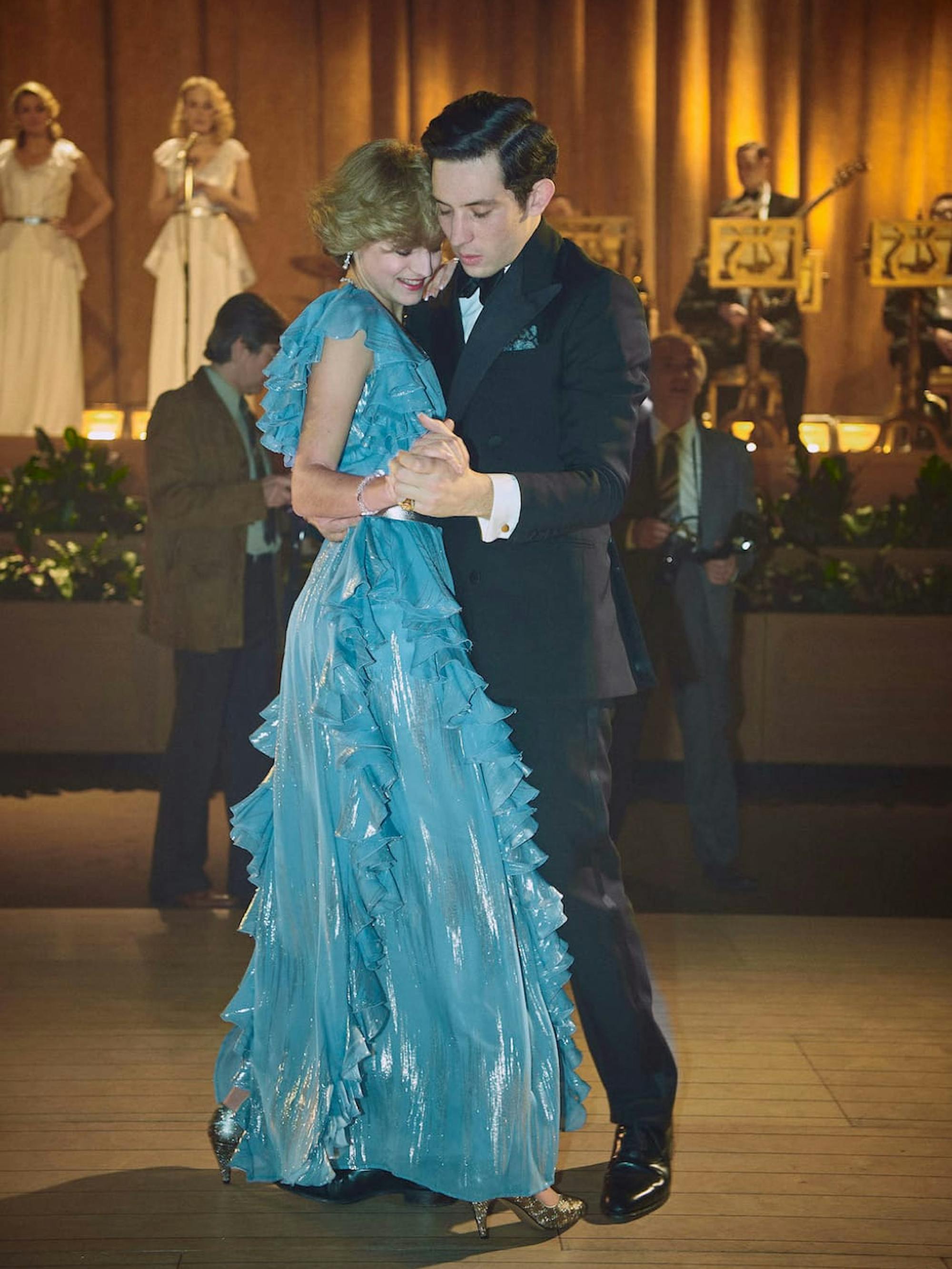 Princess Diana (Emma Corrin) and Prince Charles (Josh O’Connor) dance close together. Diana rocks a stunning, turquoise, fluttery dress and Charles wears a suit. In the background is a band and chorus.