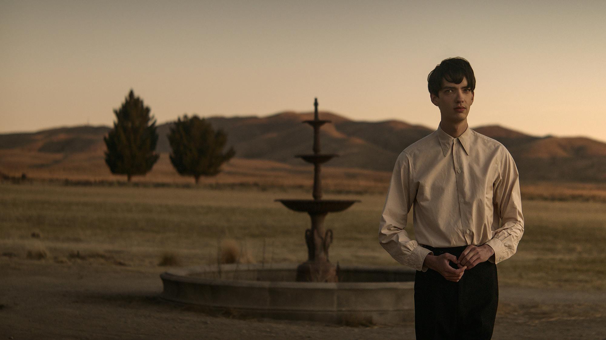 Kodi Smit-McPhee as Peter stands in front of a fountain with the striking backdrop of a mountain range and two trees standing alone in the field in front of the peaks.