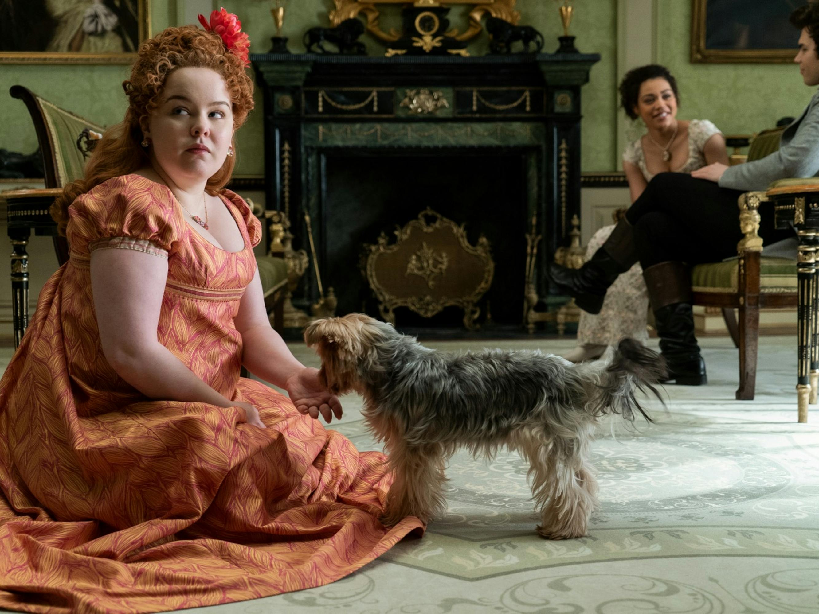 Penelope Featherington (Nicola Coughlan) wears a orange dress and orange flower, and sits on the ground to pat a dog. Her living room is ornate: there’s a fireplace, chairs, and pistachio green walls. Her cousin and a suitor sit in chairs in the distance.