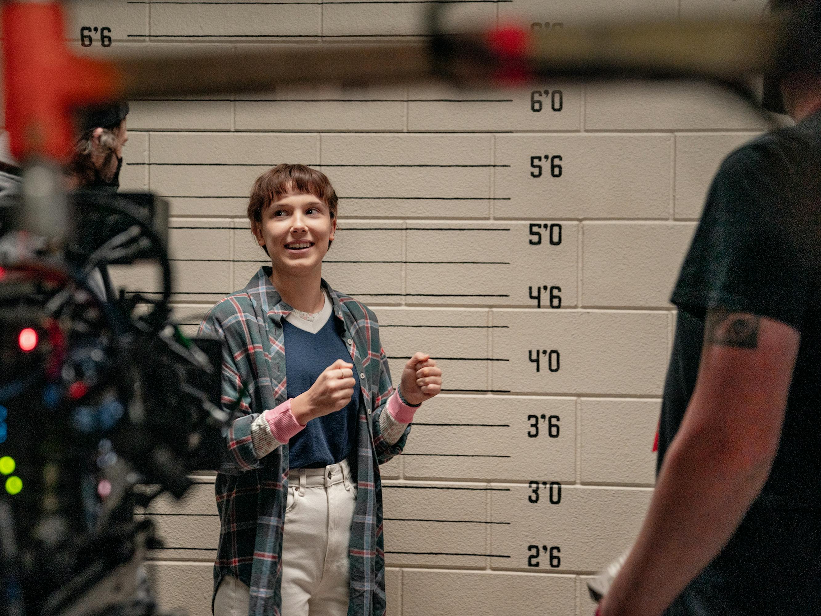 Millie Bobby Brown stands against a lined wall wearing a plaid flannel and white pants. In the foreground of this image is camera equipment.