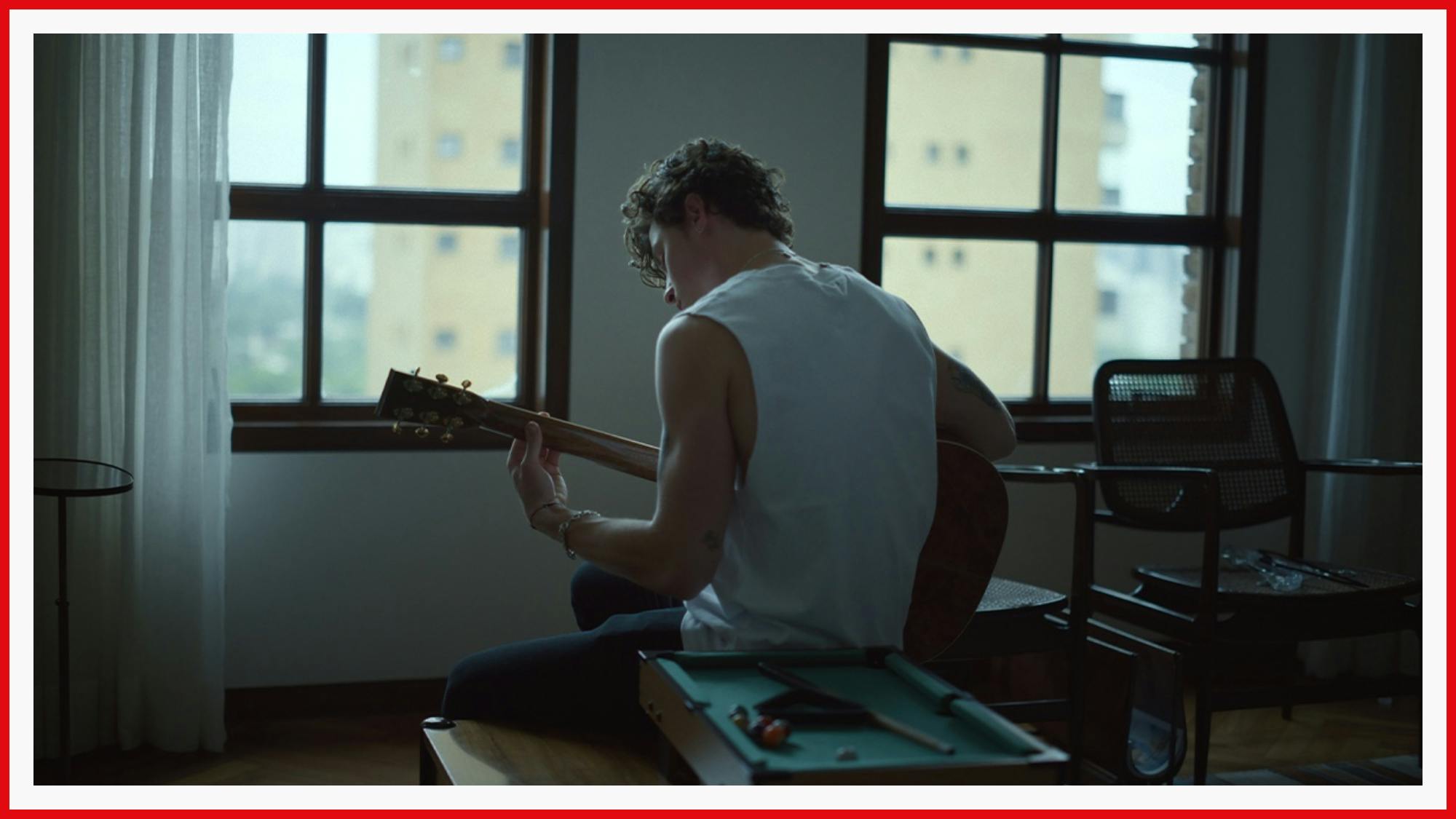 Shawn Mendes pictured from the back, wearing a white tank top, holding his guitar, probably playing something swoon-worthy. 
