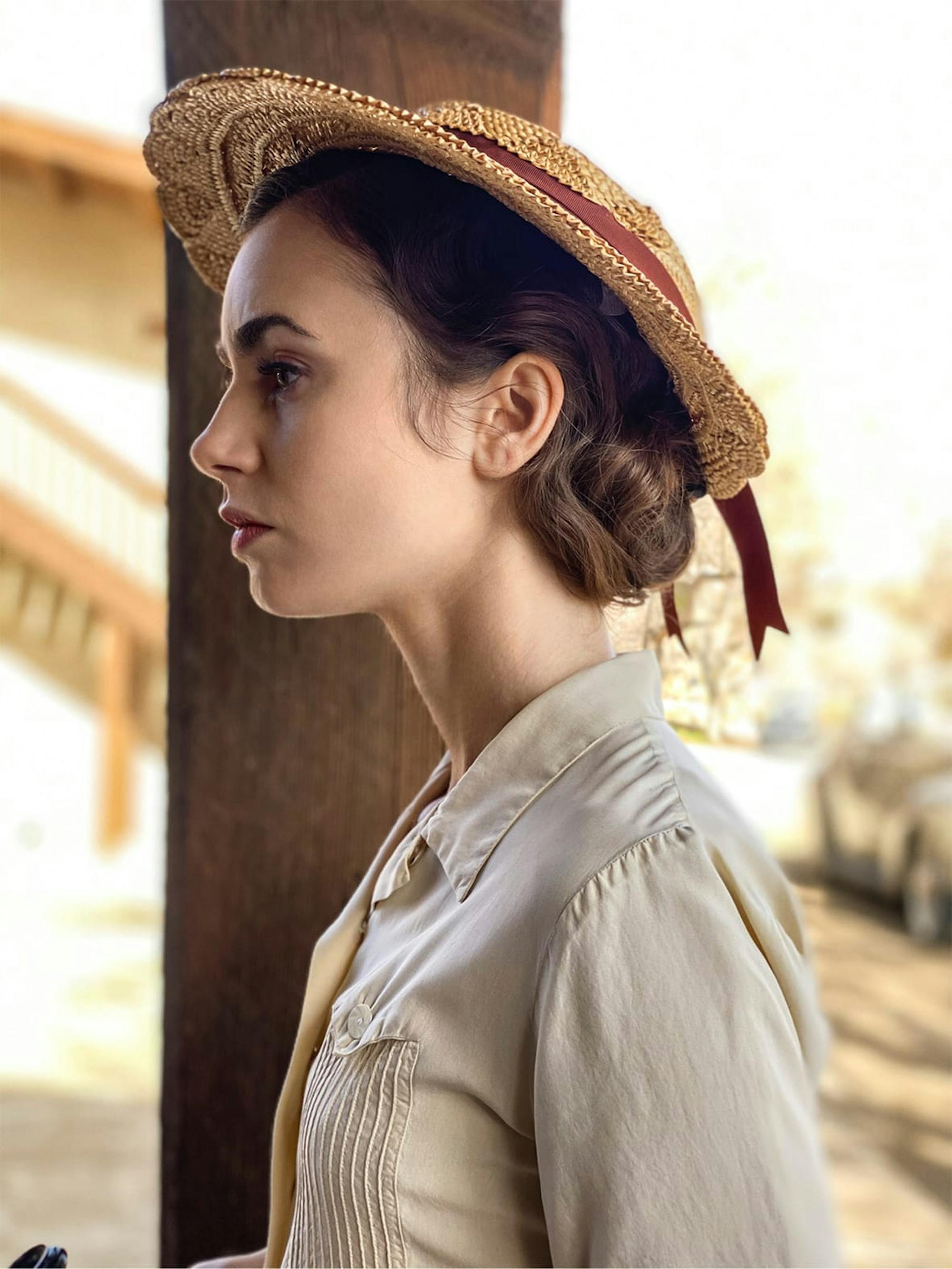 Lily Collins in character wearing a cream blouse and tan sunhat. She’s pictured in the desert light, everything browns and yellows.