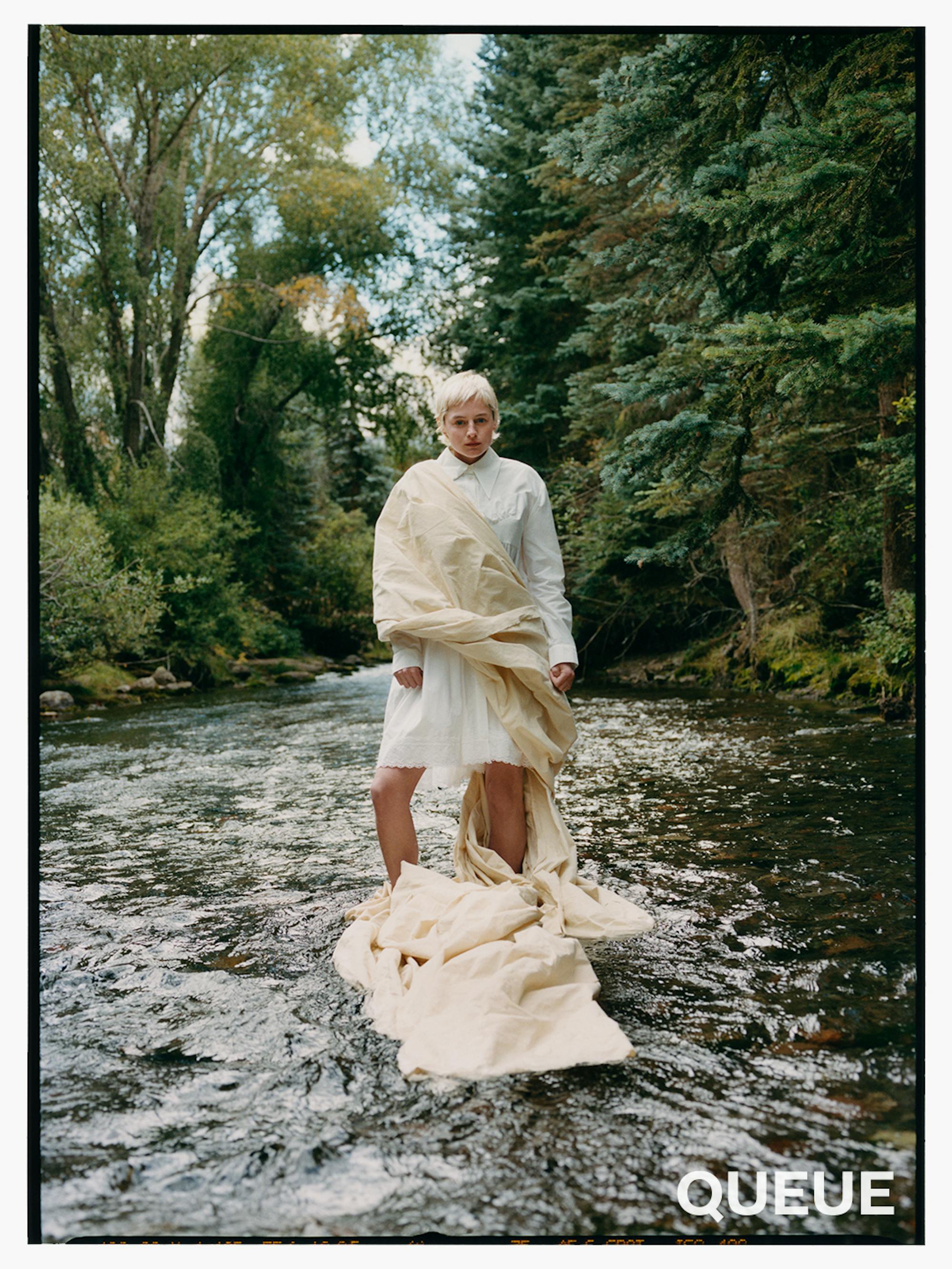 Emma Corrin wears a white toga-like outfit and stands in a running river. 