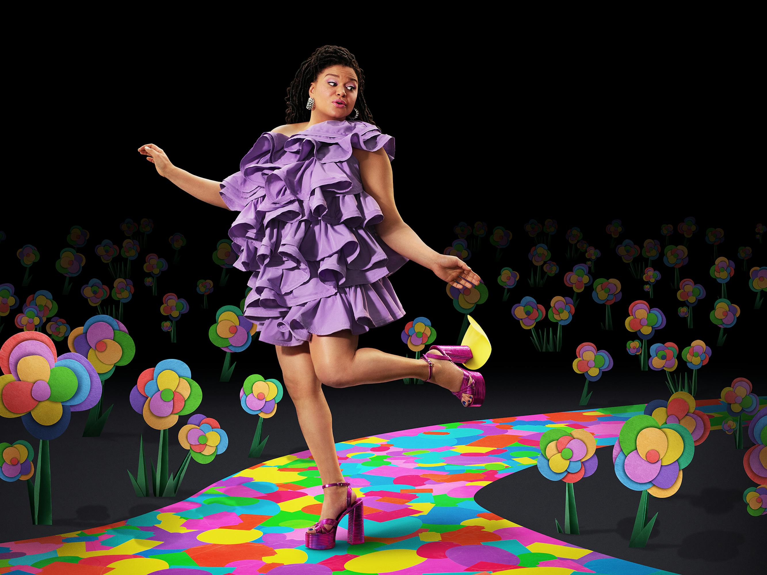 Michelle Buteau wears a ruffled purple dress and heels and walks on a sticker runway