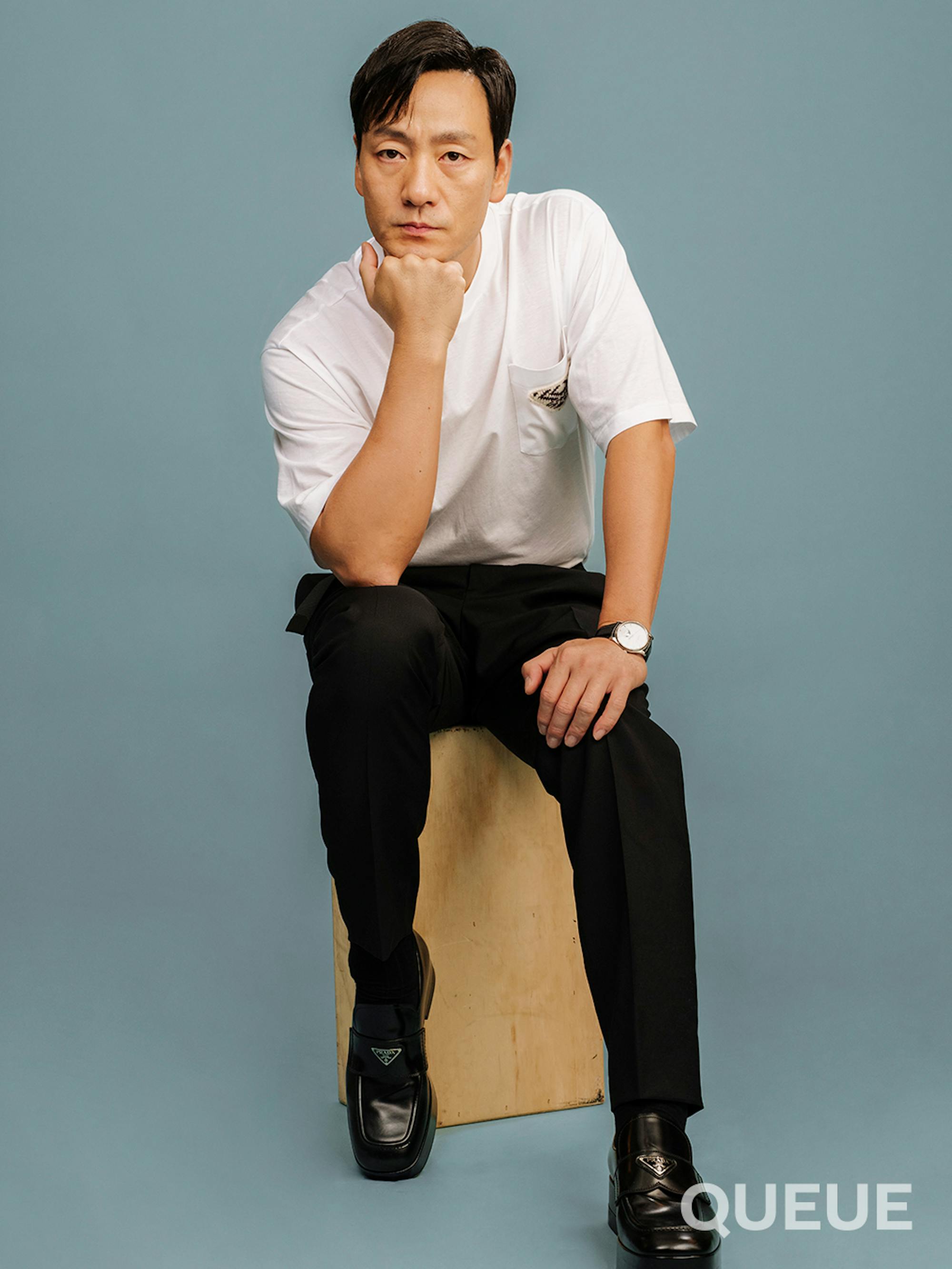 Park Hae-soo sits on an apple box in front of a gray-blue backdrop with one hand on a knee and the other under his chin. He wears a white Prada T-shirt, black trousers and black shoes.
