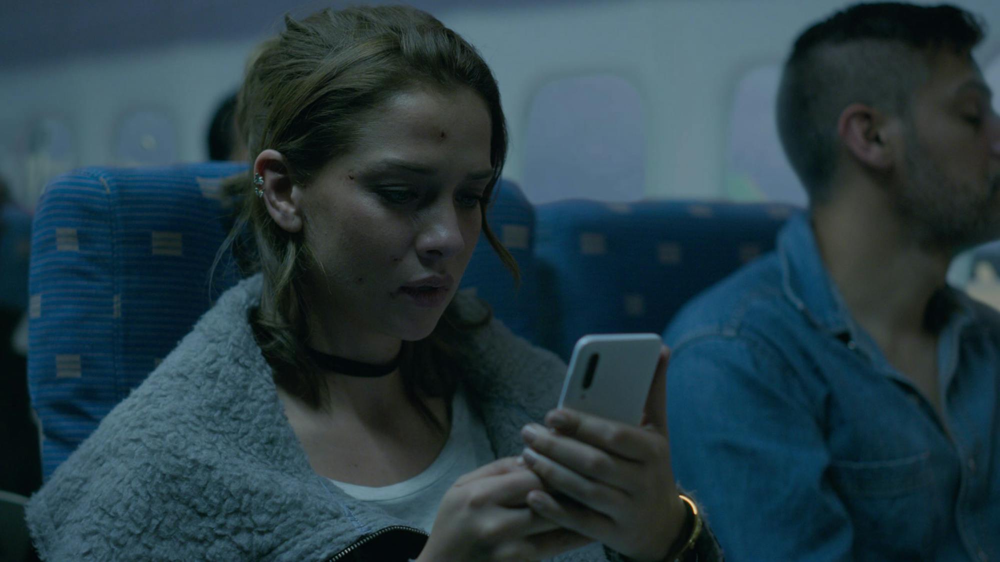 Dressed in a grey furry shawl and a black choker, Elisa (Carolina Miranda) looks at her white phone. Her face is slightly bloodied, although that doesn’t seem to concern the other passengers on the plane.