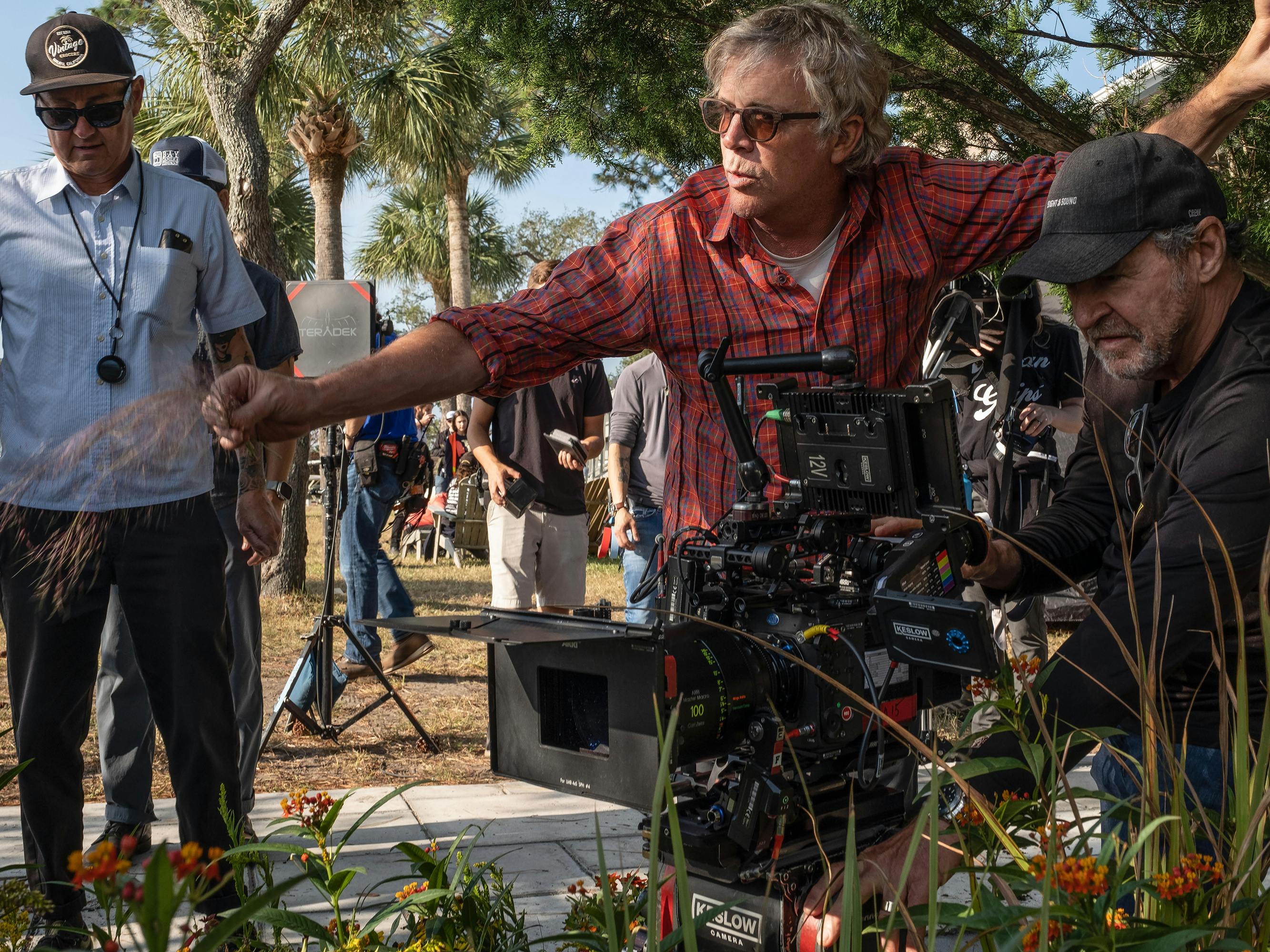 Todd Haynes behind the scenes of May December. He wears a red checkered shirt and sunglasses and talks to a man wearing all black crouching in some grass and flowers behind a massive camera.