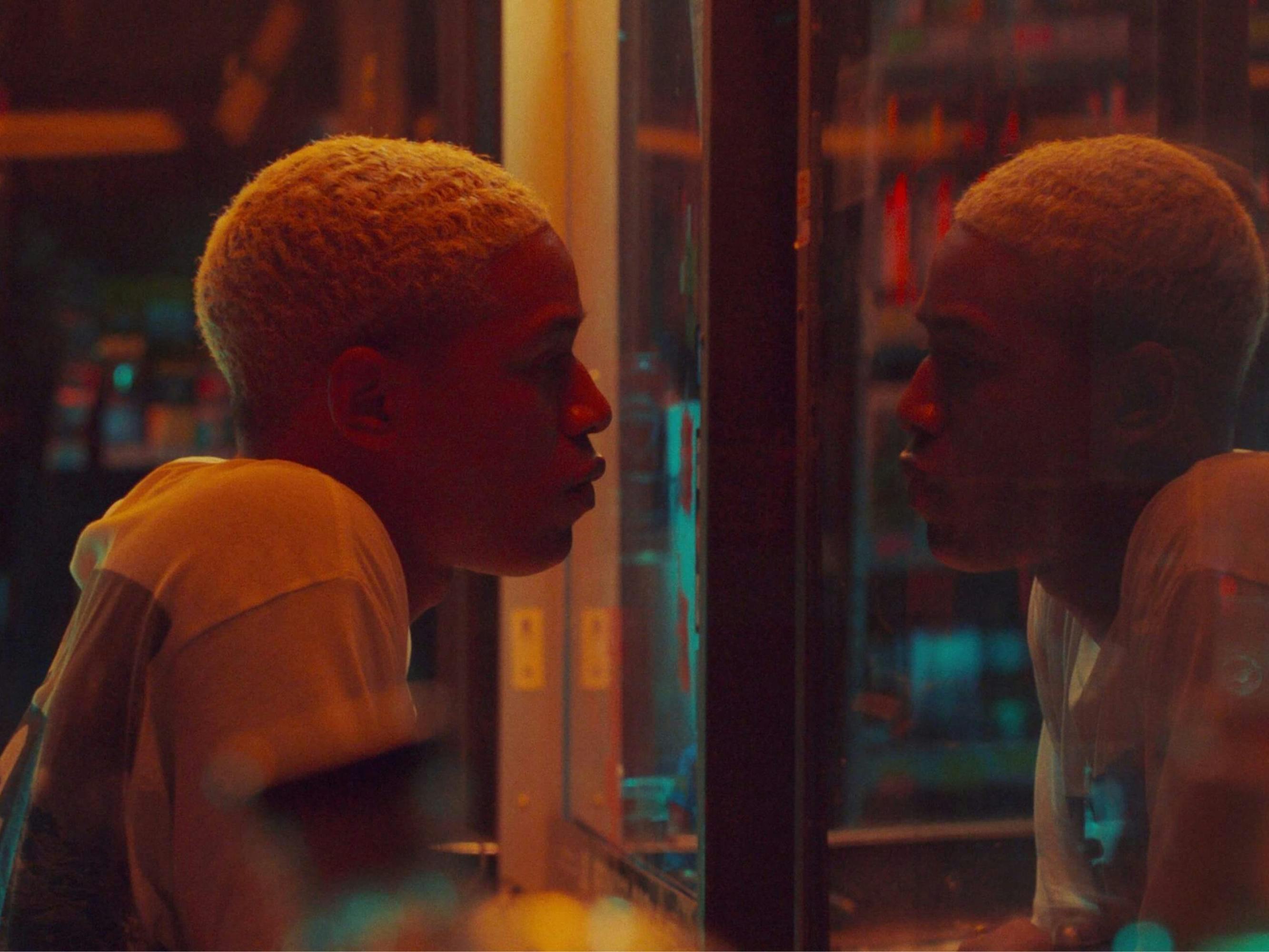 Harrison as Tyler in Waves. His hair is bleached and he wears a white t-shirt as he looks at his own reflection in the mirror. This image is made even more psychedelic by the bright flashes of neon color. 