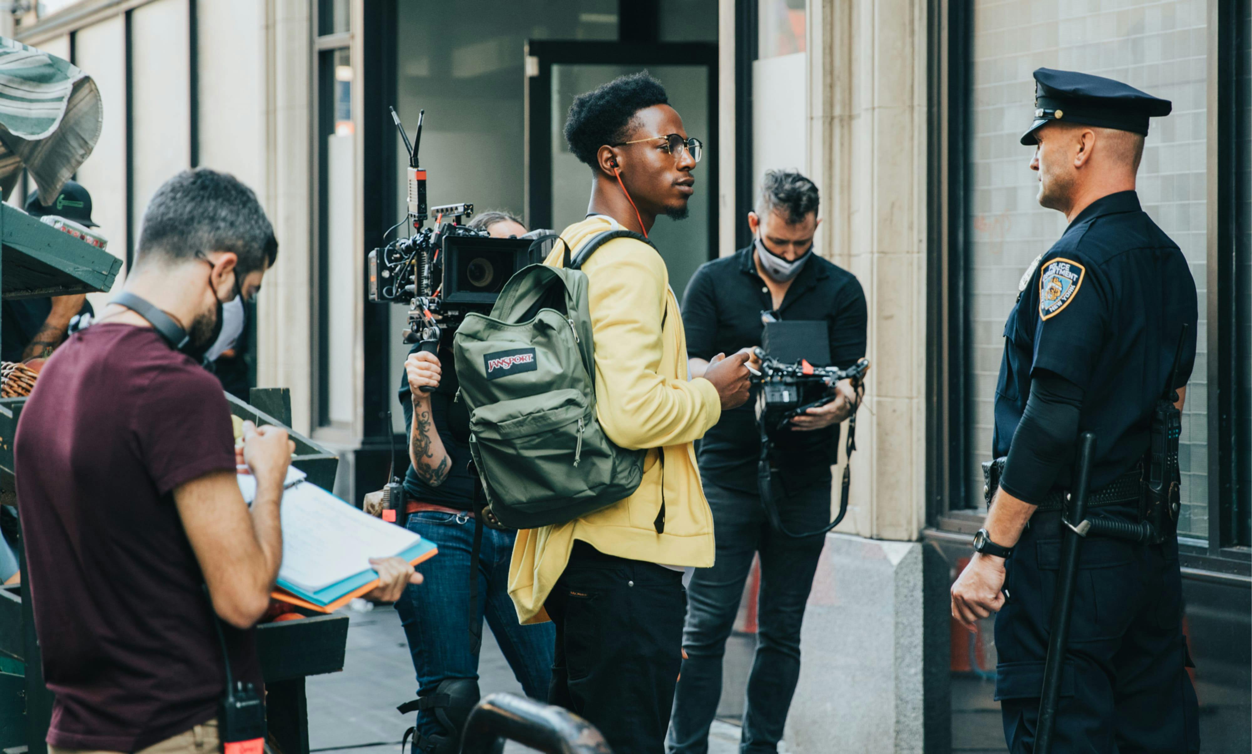 Carter (Joey Bada$$) stands in the center of this shot, flanked by cameras, cameramen, and a uniformed Merk (Andrew Howard). Bada$$ pops in a yellow hoodie, and carries a green Jansport backpack.