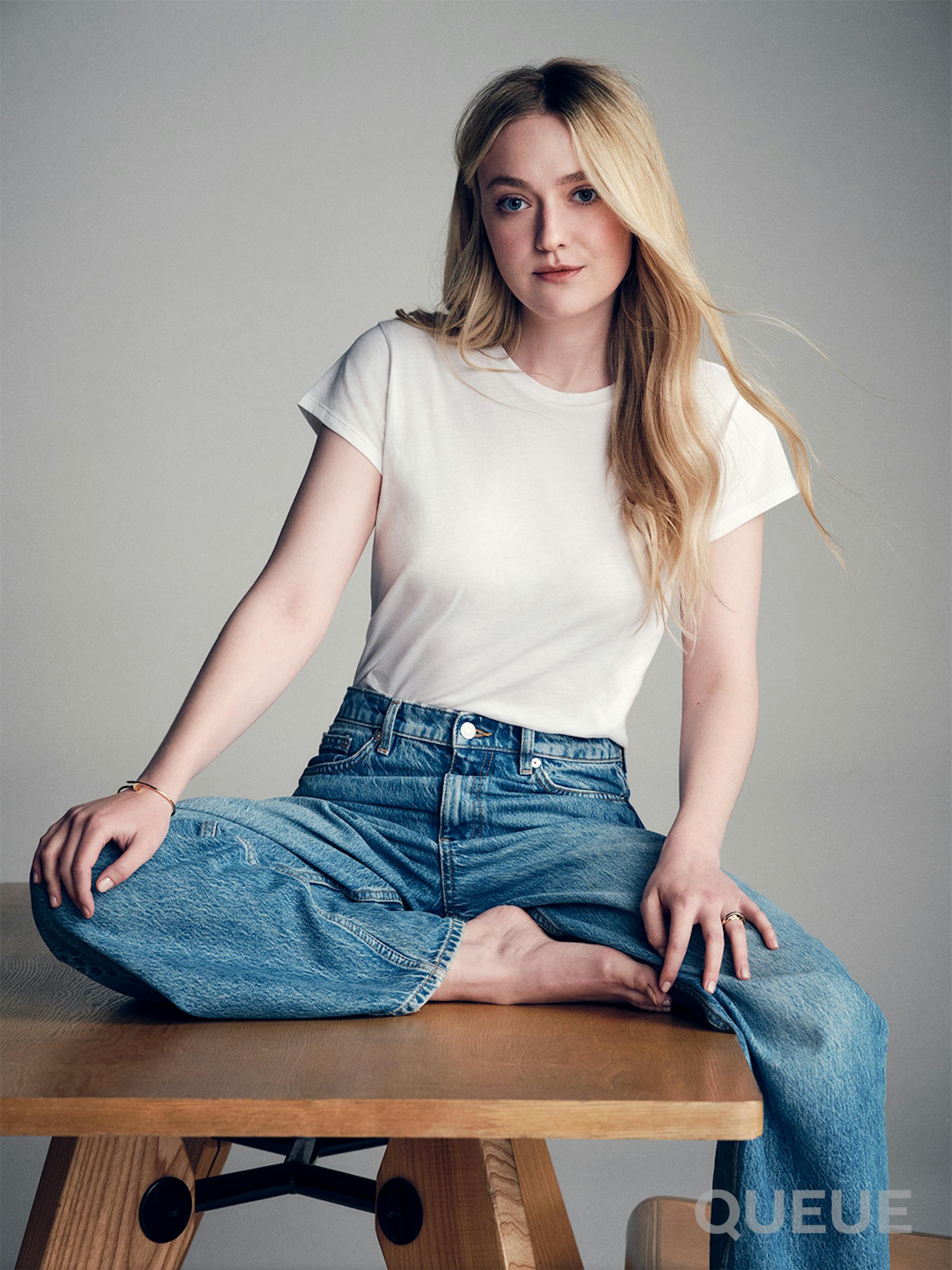 Dakota Fanning wears a white shirt and blue jeans and sits on a wooden table.