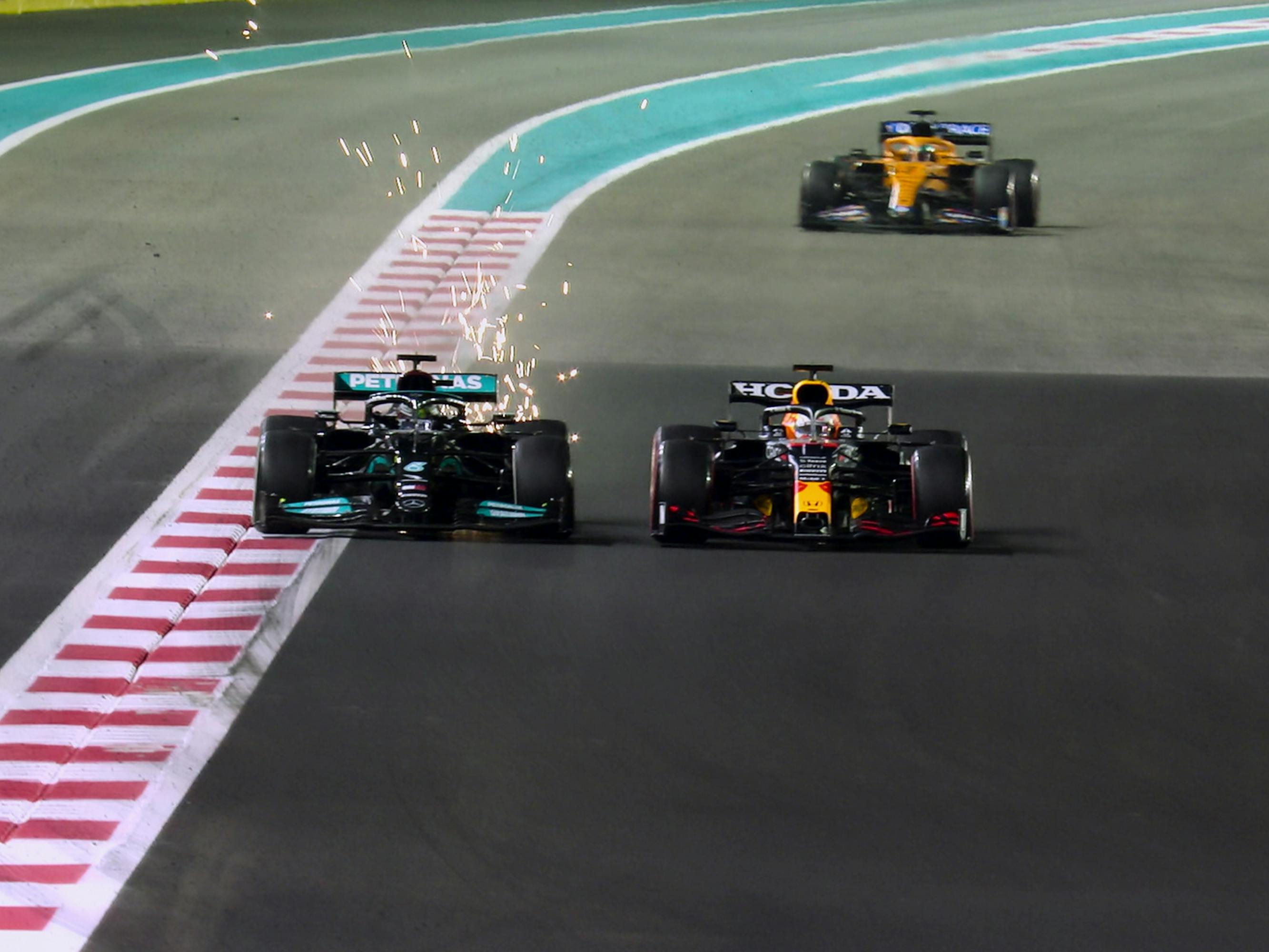 Three race cars are going down the track. Two are right next to each other with a third in the back. The front left car has sparks flying off of the back of it. 