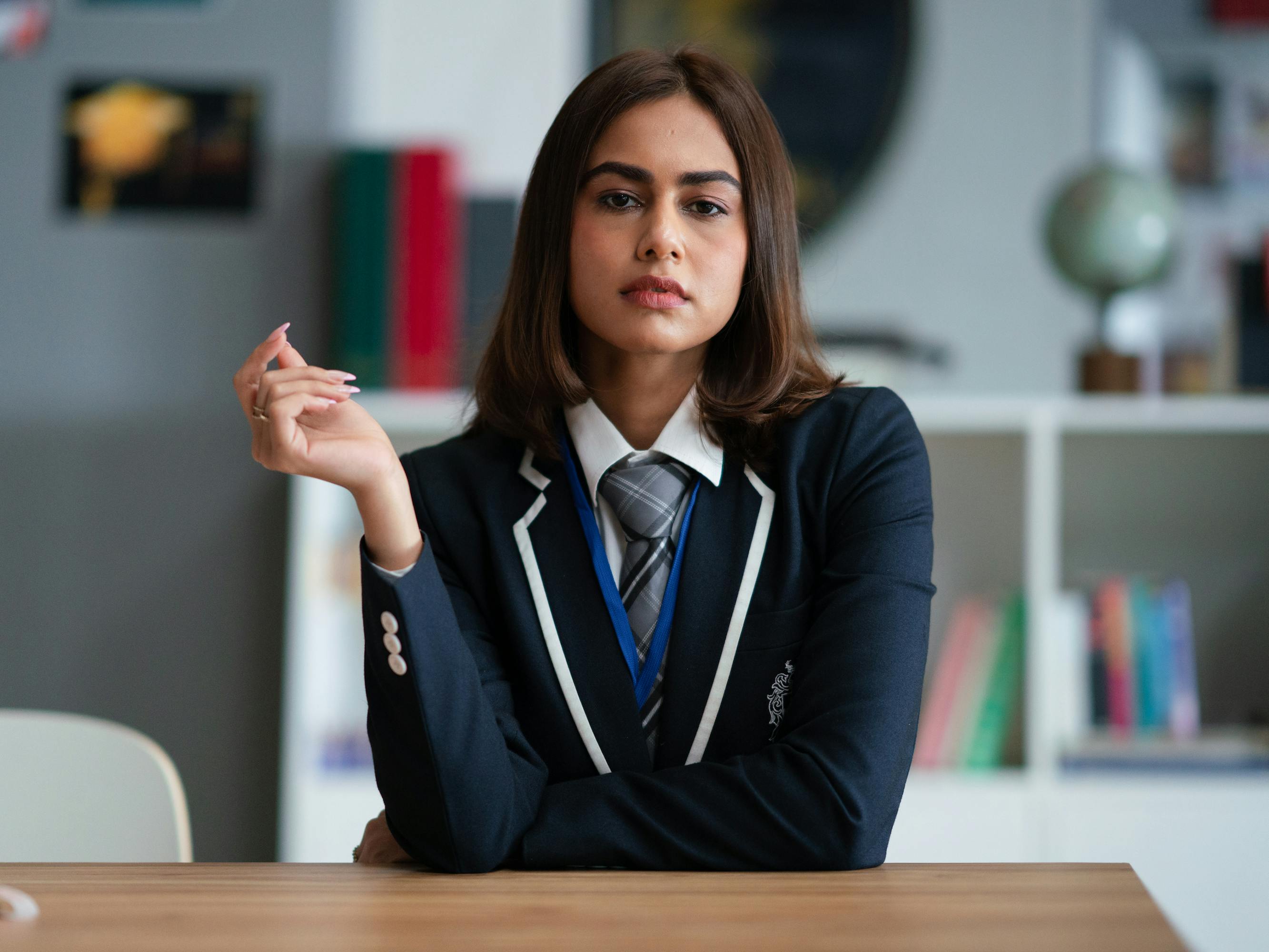 Koel (Naina Bhan) wears her school uniform: a white shirt, grey plaid tie, and navy blazer. She rests her arm on her desk and looks at the camera with attitude. 