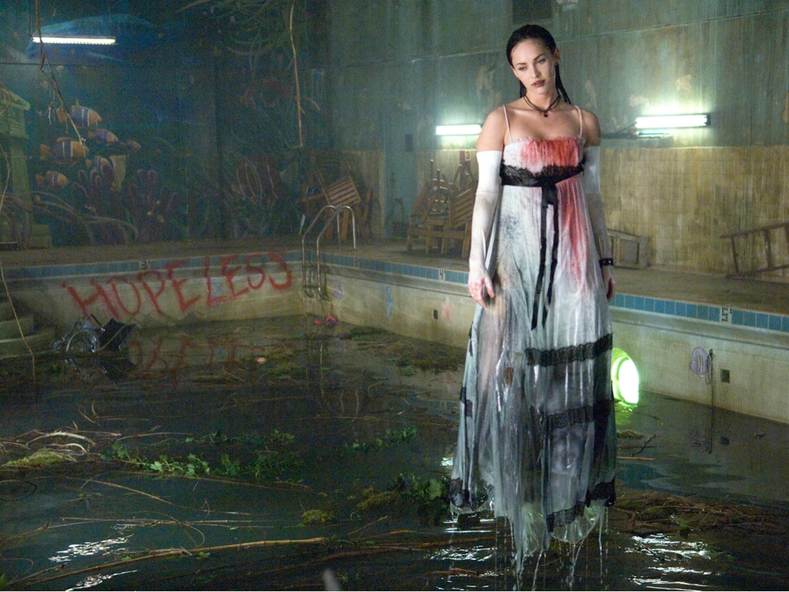 Jennifer Check (Megan Fox) in Jennifer’s Body. She wears a white dress which is streaked with black and red, and stands in a pool cluttered with branches and leaves.