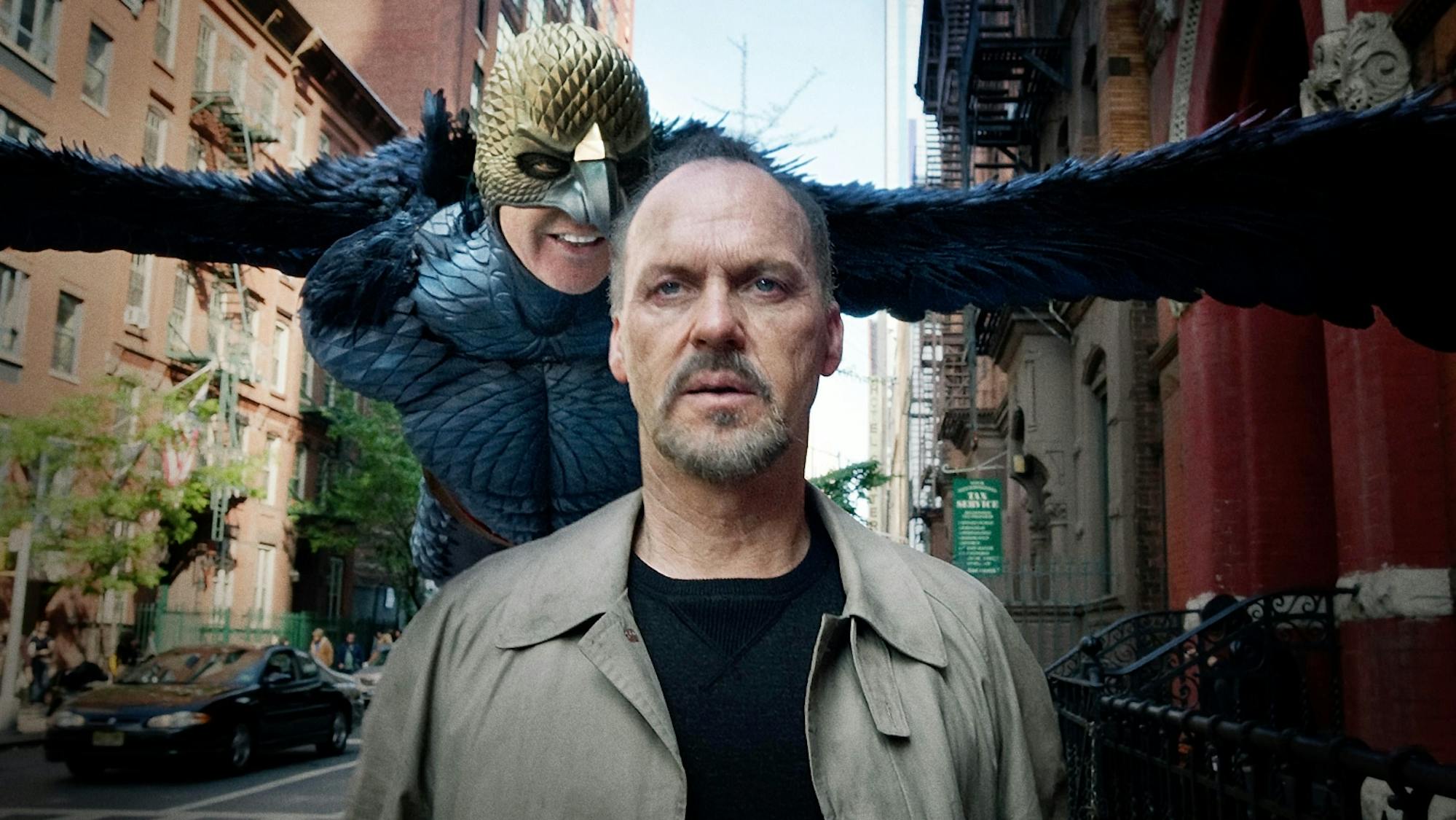 Riggan Thomson (Michael Keaton) in Birdman wears an olive colored jacket and black t-shirt. Behind him is the black costumed birdman. They make their way down a New York City street.