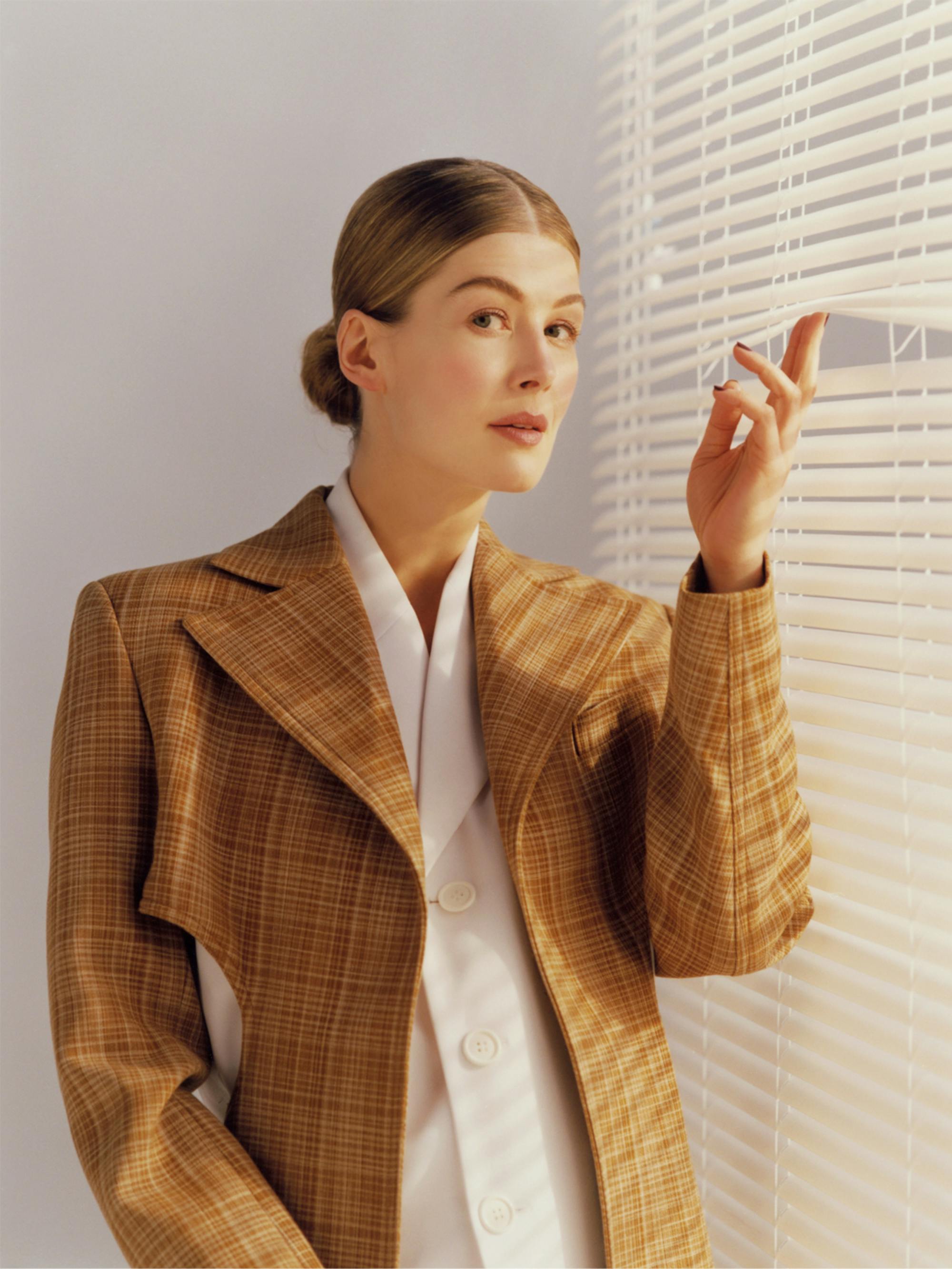 Rosamund Pike poses in a checkered jacket and white shirt. She looks askance at the camera as she uses two fingers to lift up a panel on a set of venetian blinds.