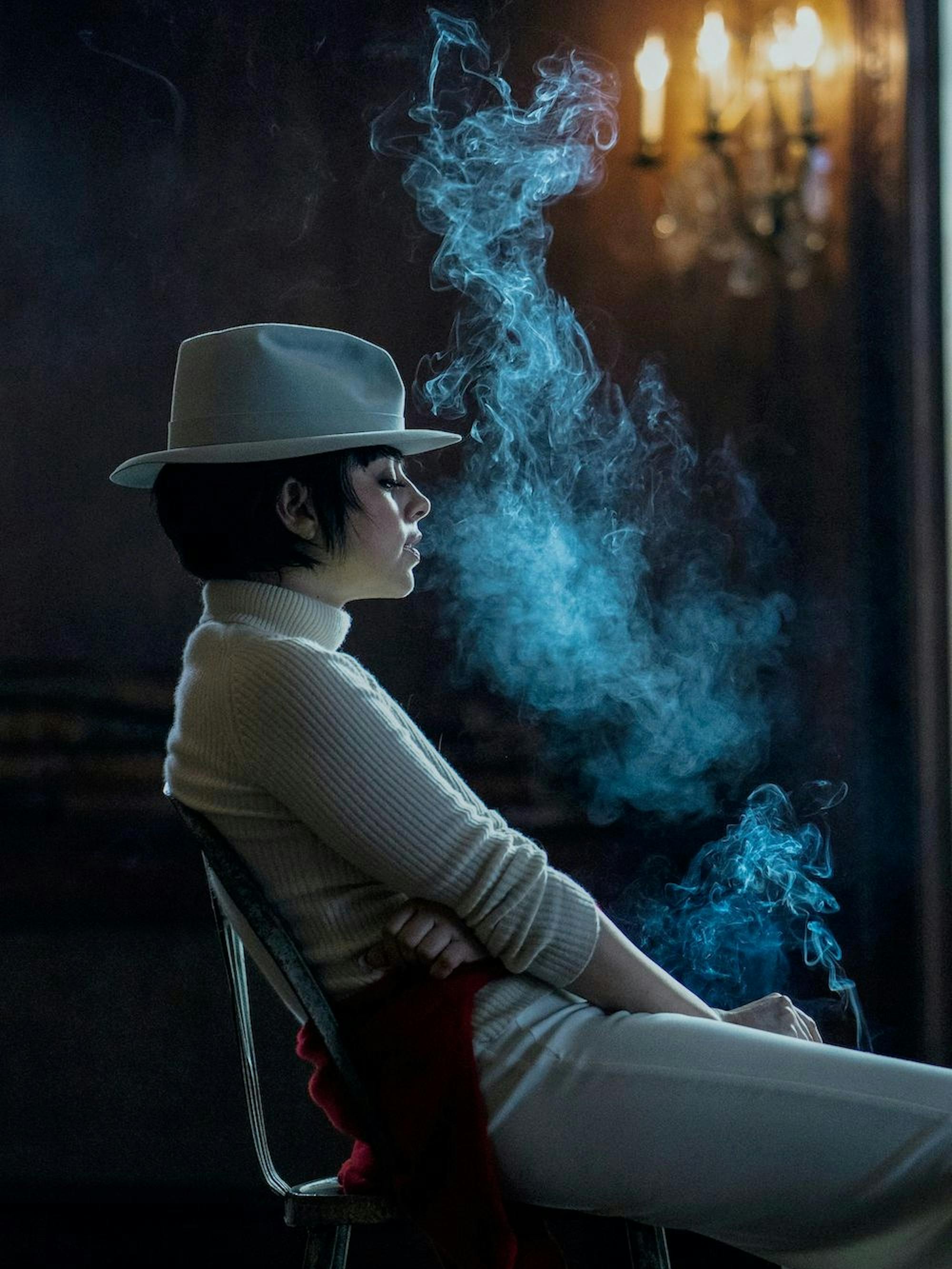 Liza Minnelli (Krysta Rodriguez) leans back in a chair, shrouded by a cloud of her cigarette smoke. She is dressed in all white, included fedora, and the eerie shot is lit only by a light fixture in the background.