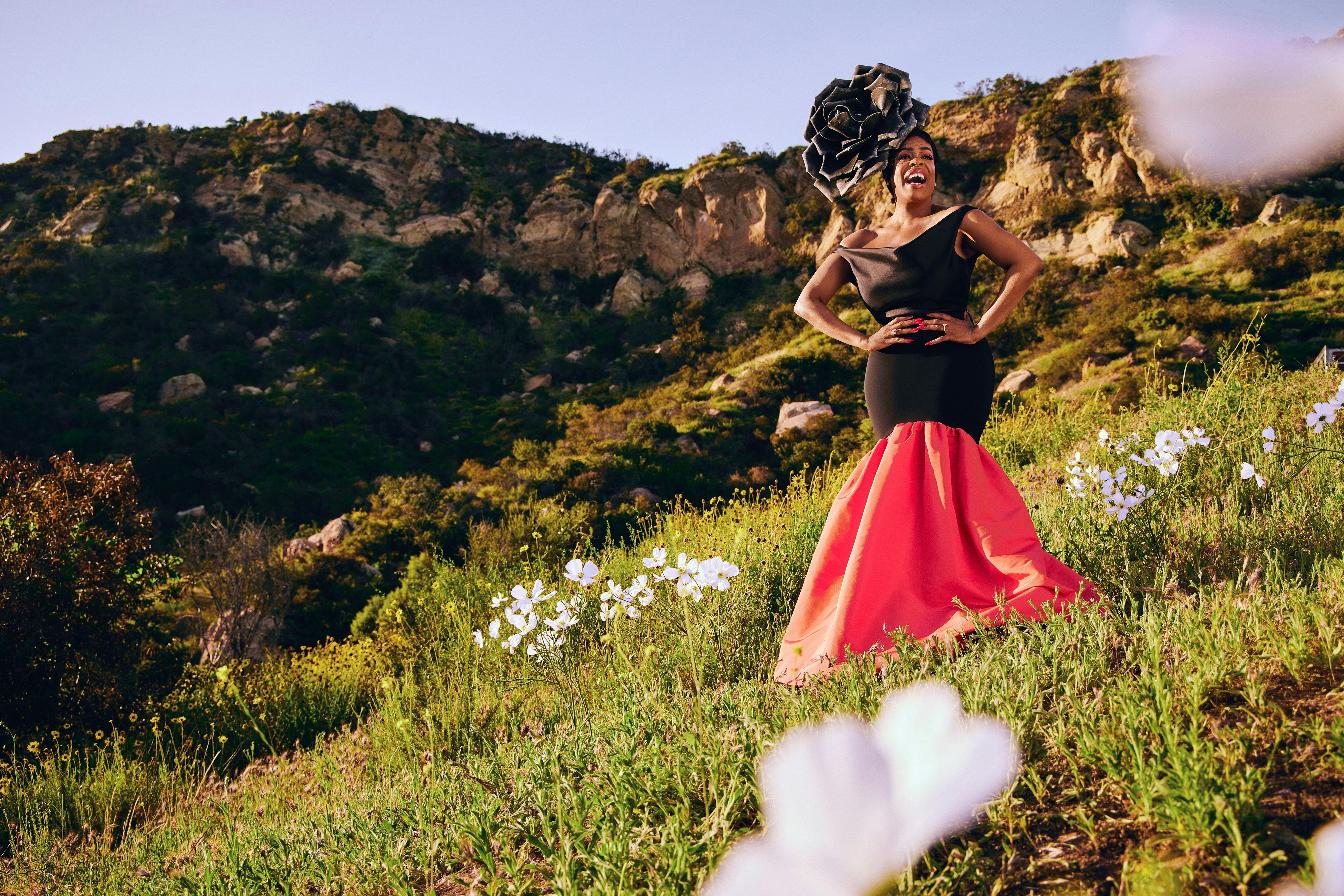 Niecy Nash-Betts wears a red-and-black dress and an elaborate black headpiece and stands on a grassy hillside, dotted with white flowers. 
