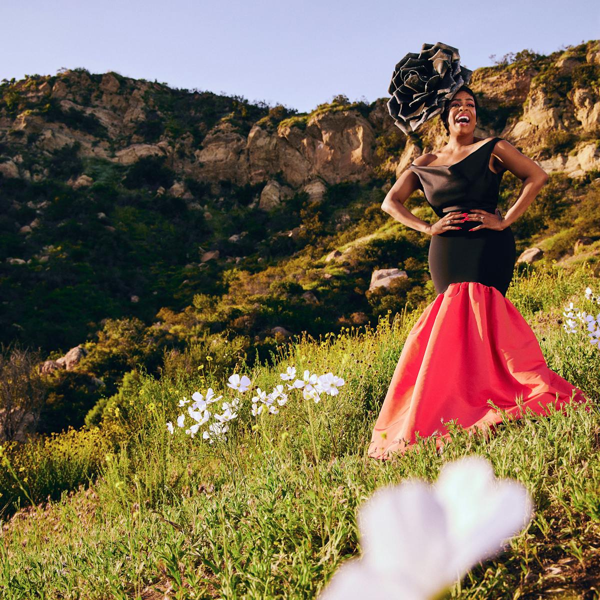 Niecy Nash-Betts wears a red-and-black dress and an elaborate black headpiece and stands on a grassy hillside, dotted with white flowers. 