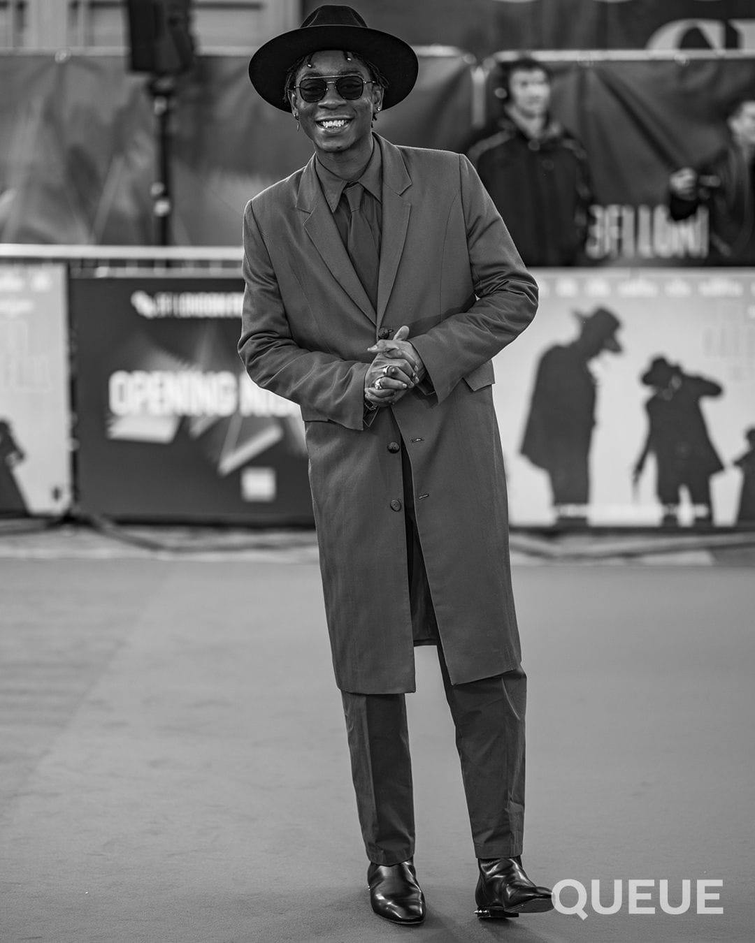 RJ Cyler in a wide brimmed hat, sunglasses, and knee-length coat on the red carpet