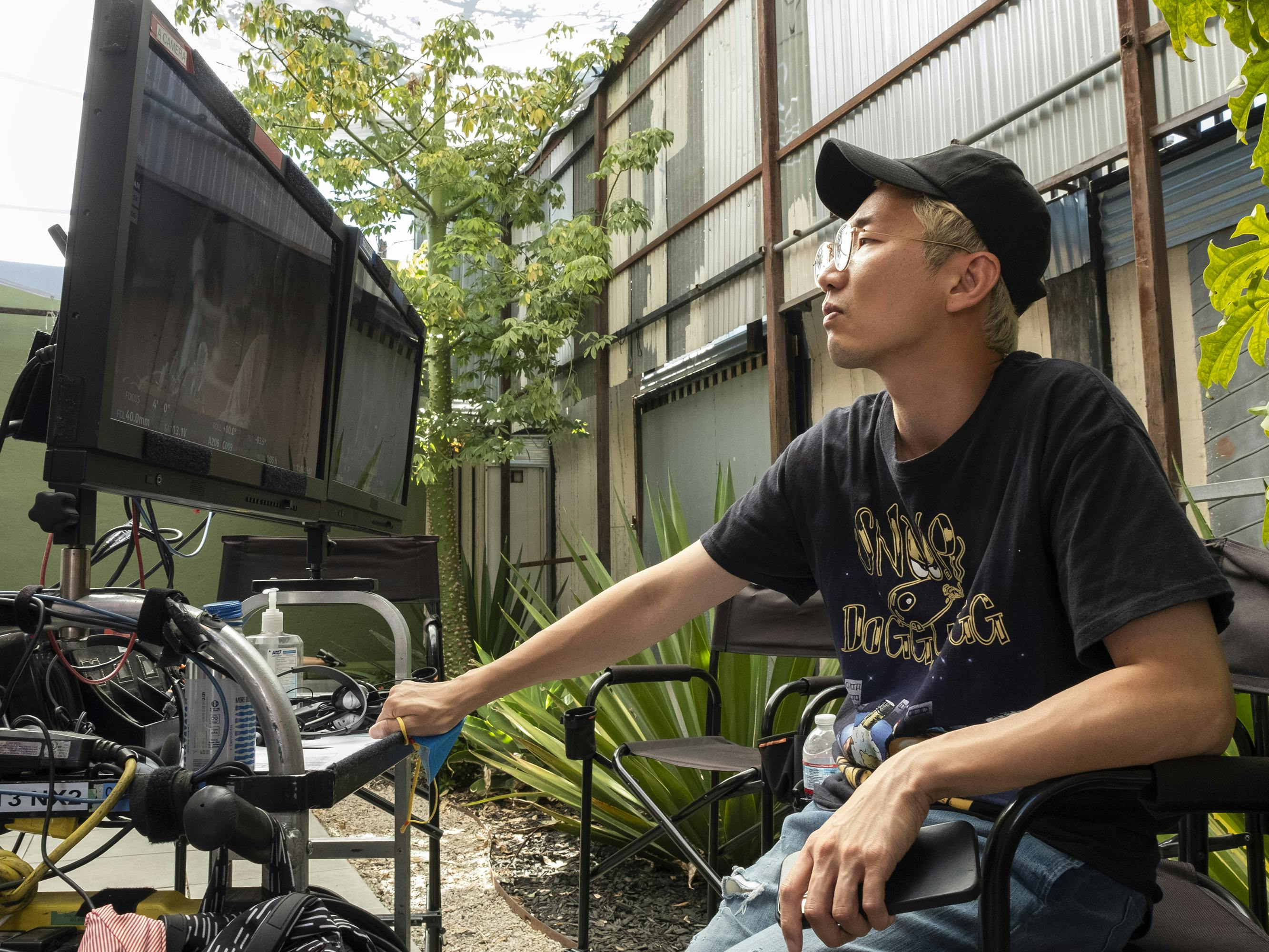 Lee Sung Jin wears all black — including a black baseball cap — and sits behind a monitor in a plant-filled outdoor space.