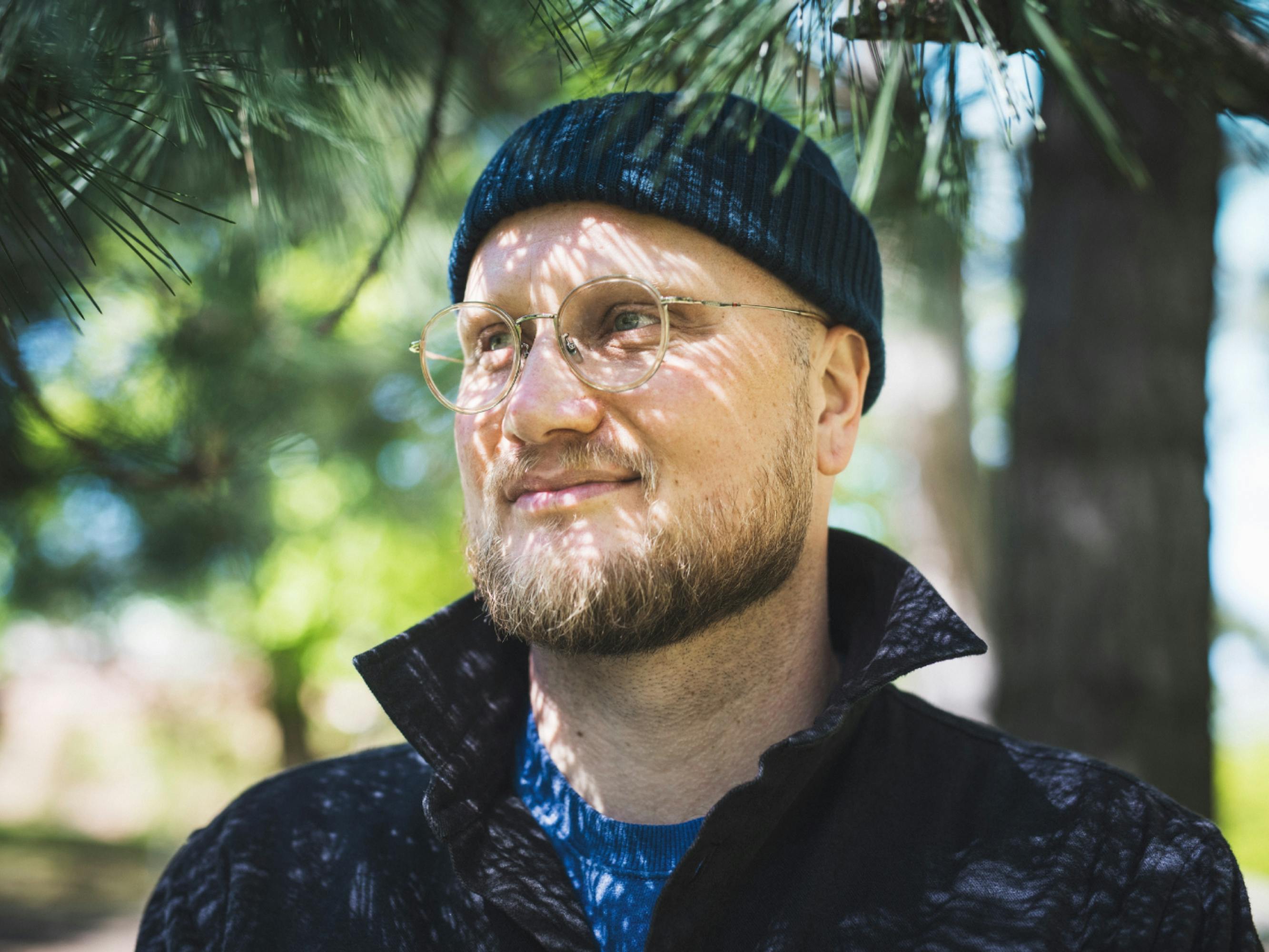 Co-creator and head writer Oskar Söderlund against a brightly lit background filled with greenery. He wears a black beanie, circular glasses, and a black fleece.