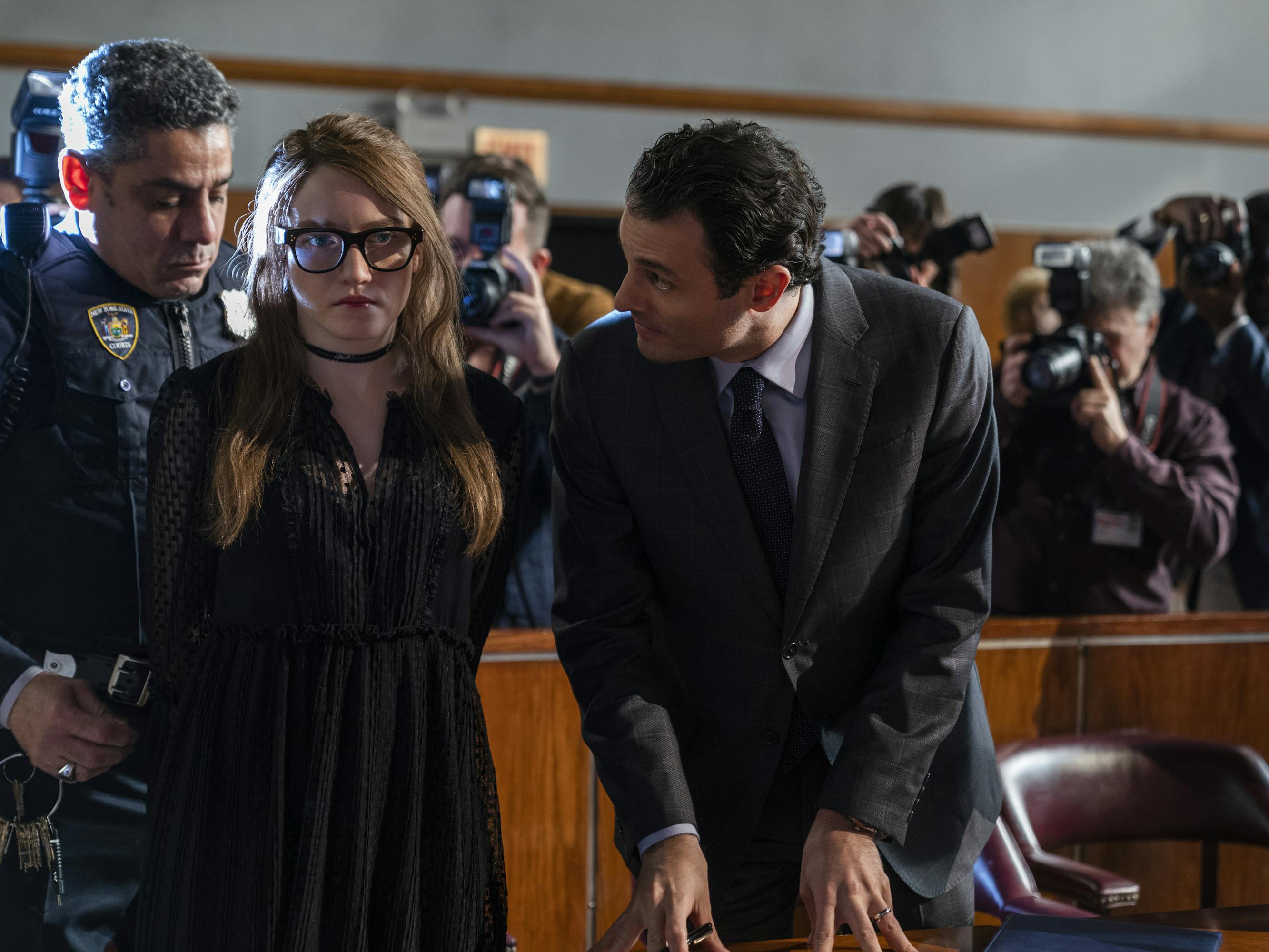 Julia Garner and Arian Moayed stand together in a courtroom as Garner gets handcuffed by a police officer. Garner wears a black dress, glasses, and a choker. Moayed wears a dark suit.