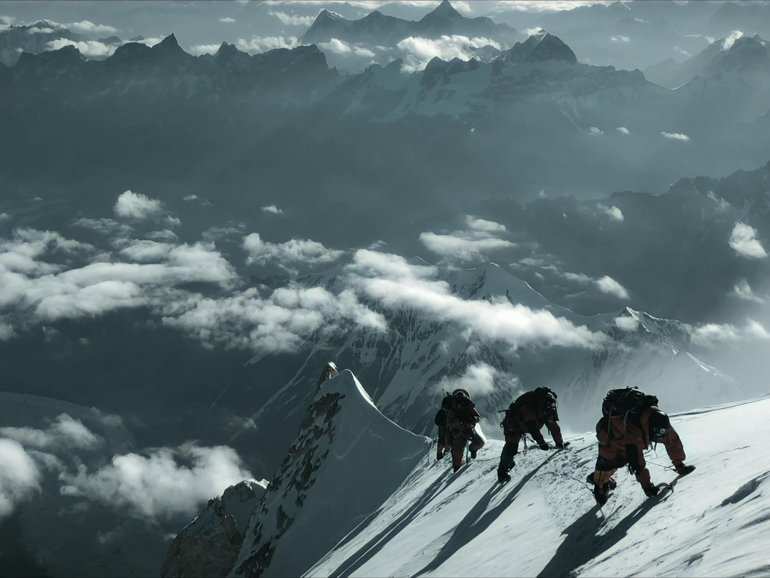 Hikers straddle a rope up a perilous, cloudy, snowy ledge.