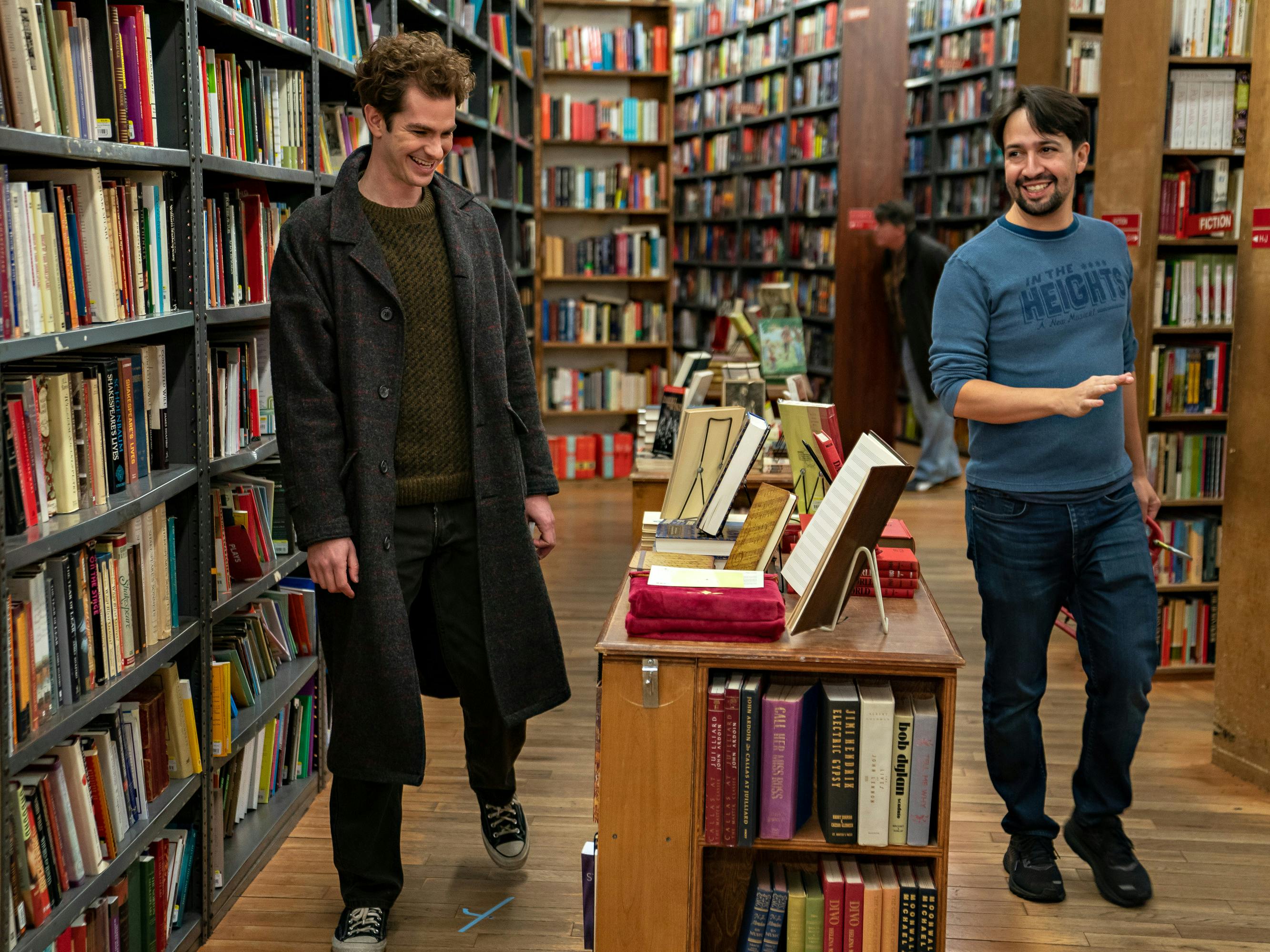 Andrew Garfield and Lin-Manuel Miranda laugh in a bookstore. Garfield wears a long black coat, and Miranda wears a teal shirt and jeans.