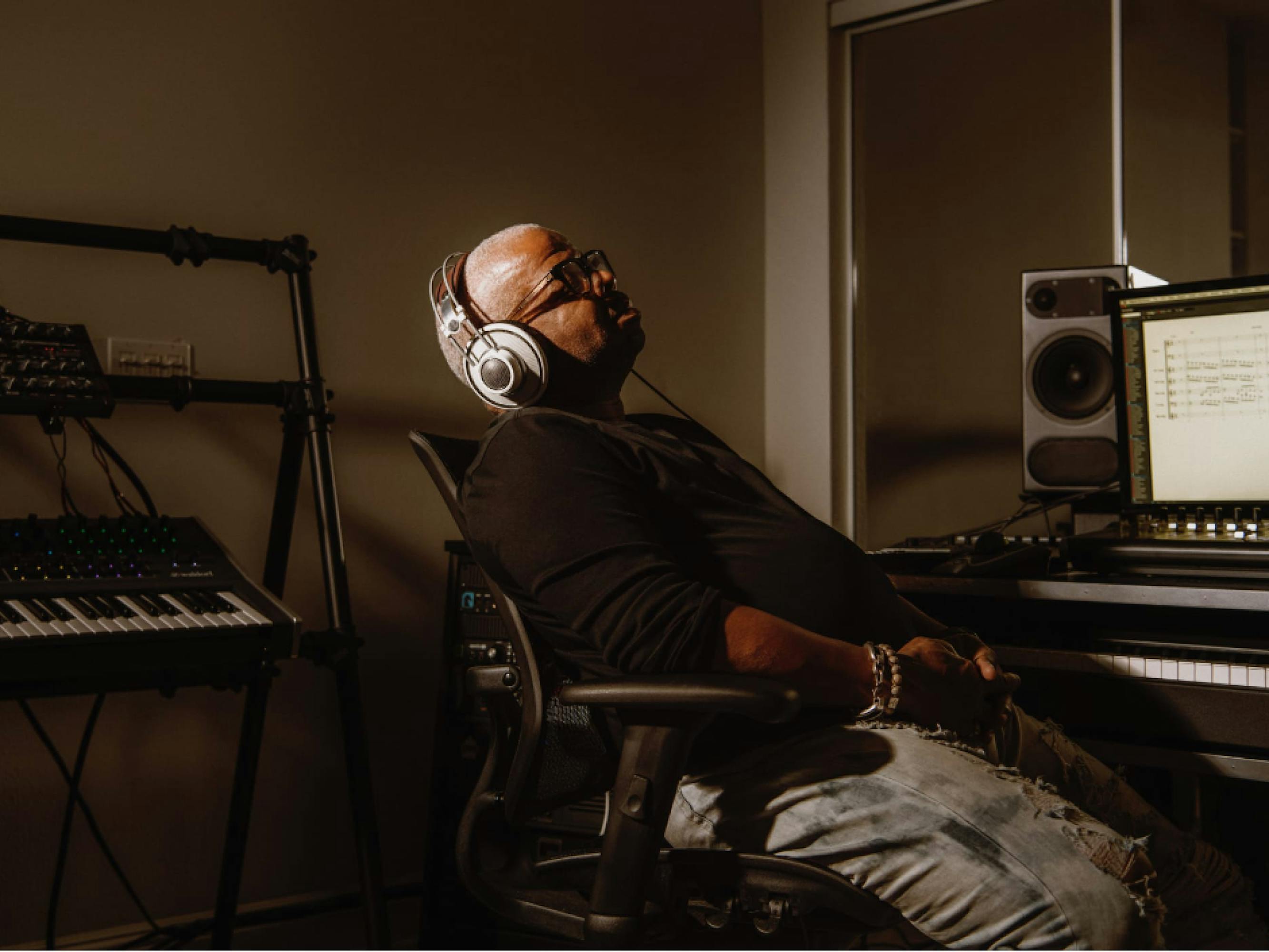 A photograph of Terence Blanchard in the studio. He is seated at a desk with a computer and speakers on it. He leans back in his chair, eyes closed, a pair of headphones over his ears. He wears a black shirt and ripped blue jeans. Behind him, we can see a keyboard. 