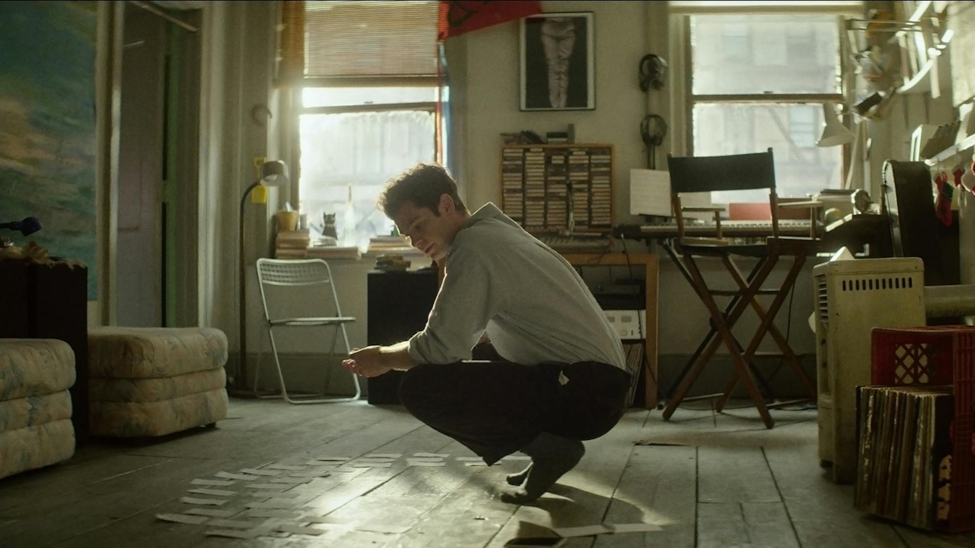 Andrew Garfield as Jonathan Larson wears a light shirt and dark pants as he squats in his apartment in front of slips of papers.  Behind him is a desk, milk crates, cushions, books, and windows.