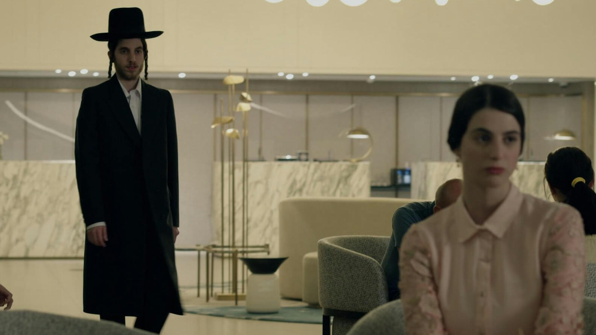 A man in a black coat and hat stands in the background looking at a woman in a pink blouse in the foreground, who seems unaware of his presence. The room is a hotel lobby and is scattered with small grey chairs, marble, and light fixtures.